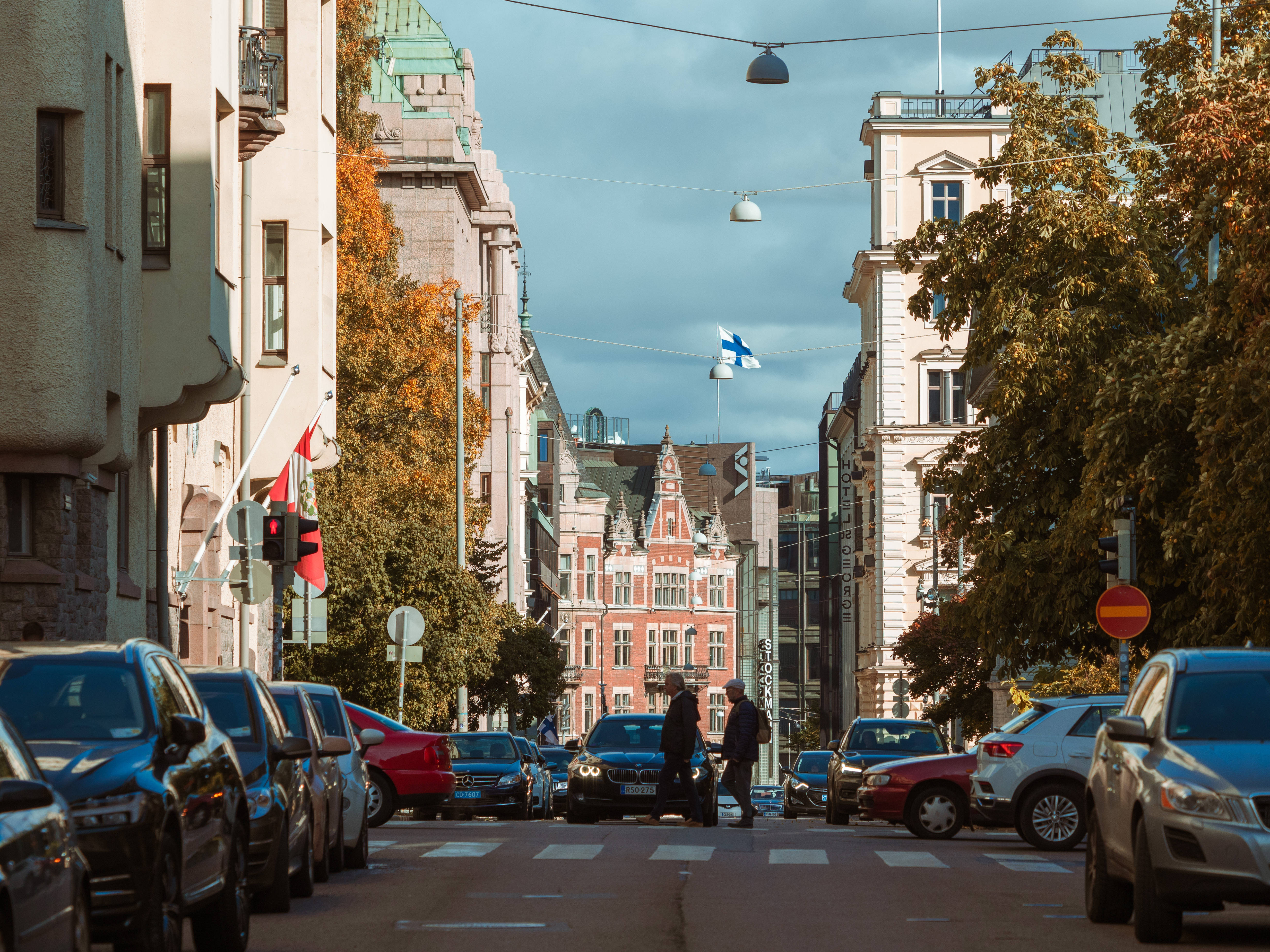 Finland 桌布 壁紙 — 一條陽光明媚的歐洲街道，兩旁是歷史建築和秋樹，行人正在停泊的汽車附近過馬路。