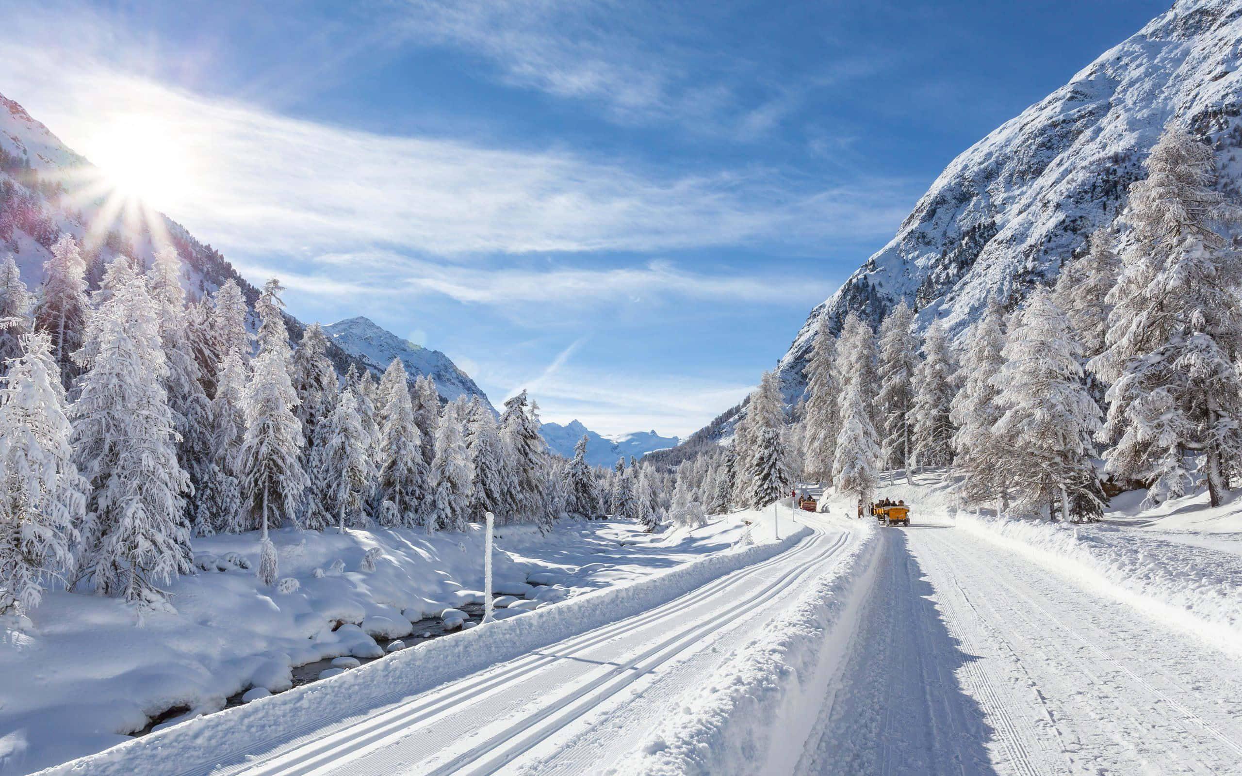 Icy Road 桌布 壁紙 — 一條陽光明媚的積雪山路蜿蜒穿過白雪覆蓋的松林，天空湛藍，雲朵輕柔。