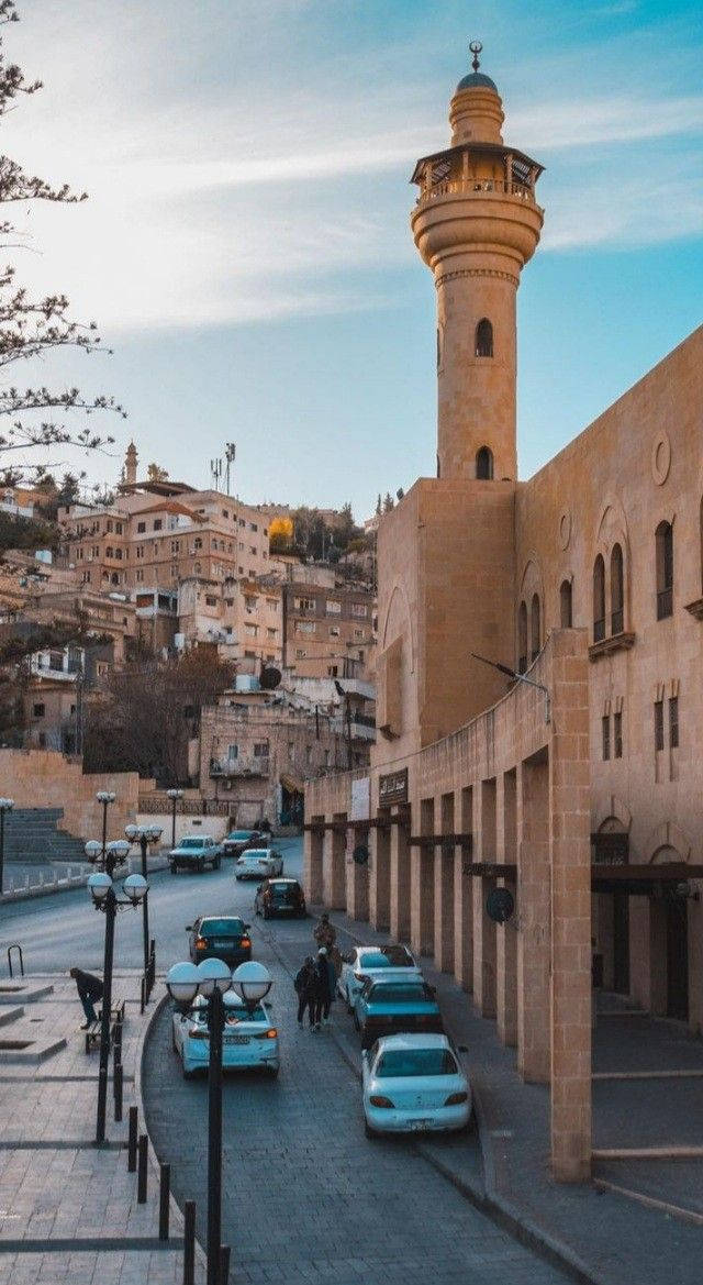 Jordan 桌布 壁紙 — 一條陽光明媚的街道，位於歷史悠久的中東小鎮，高聳的清真寺宣禮塔矗立在陶土建築和停放的汽車之上。
