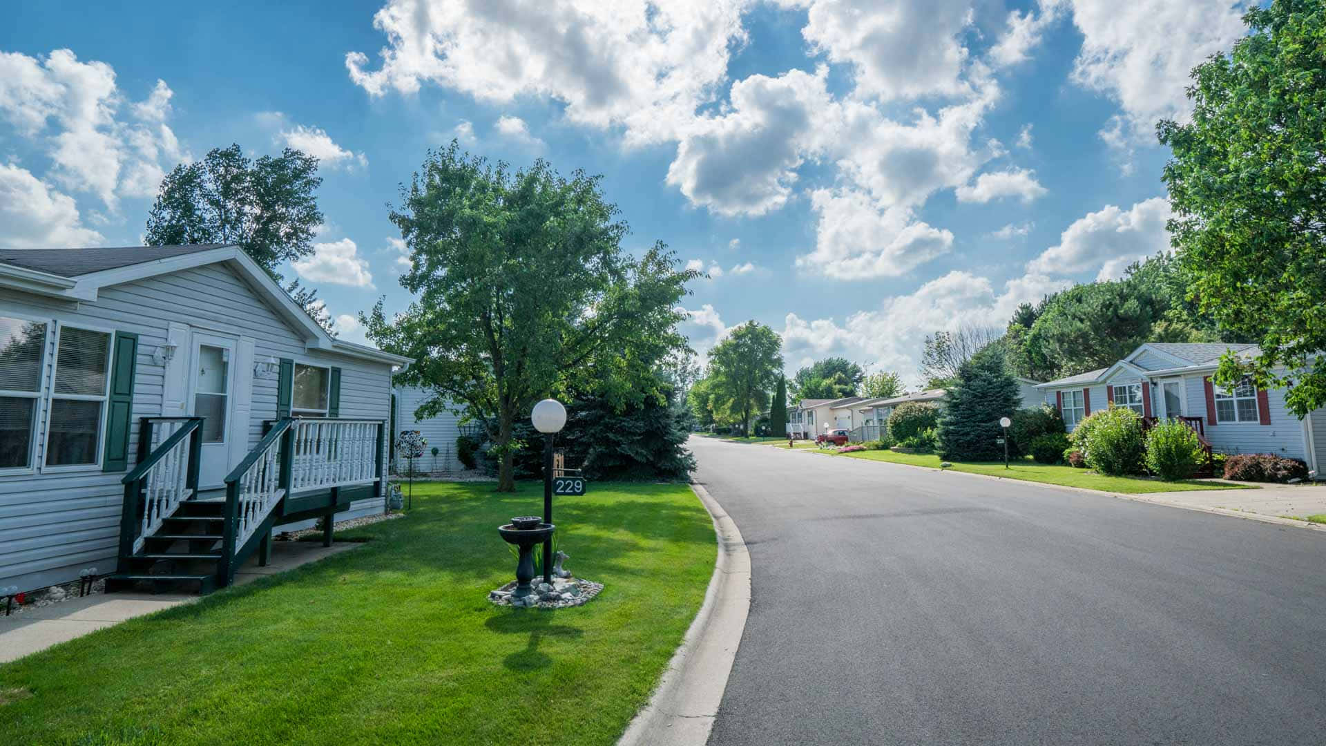 Neighborhood 桌布 壁紙 — 一條陽光明媚的郊區街道，兩旁是白色的移動房屋和茂密的綠色草坪，上方是明亮的藍天和蓬鬆的白雲。