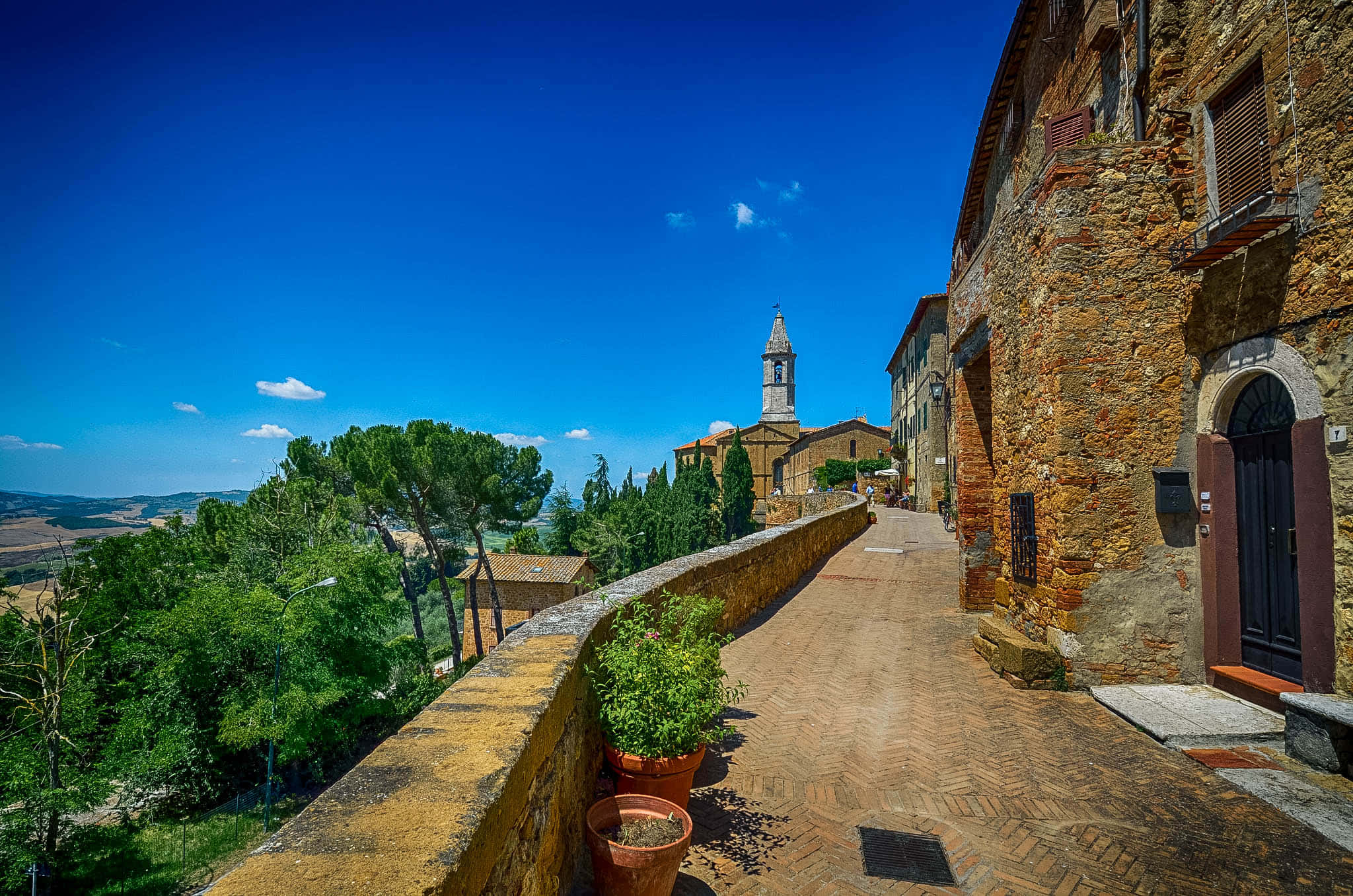 Tuscany 桌布 壁紙 — 一條陽光明媚的鵝卵石小徑蜿蜒經過古樸的石頭建築，通往一座教堂塔樓，俯瞰著晴朗藍天下鬱鬱蔥蔥的綠色山丘。