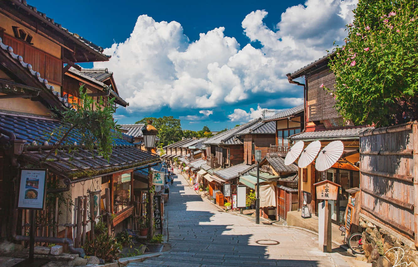 日本 桌布 壁紙 — 一條陽光明媚的鵝卵石街道，兩旁是傳統木建築和紙傘，上方是明亮的藍天和蓬鬆的白雲。
