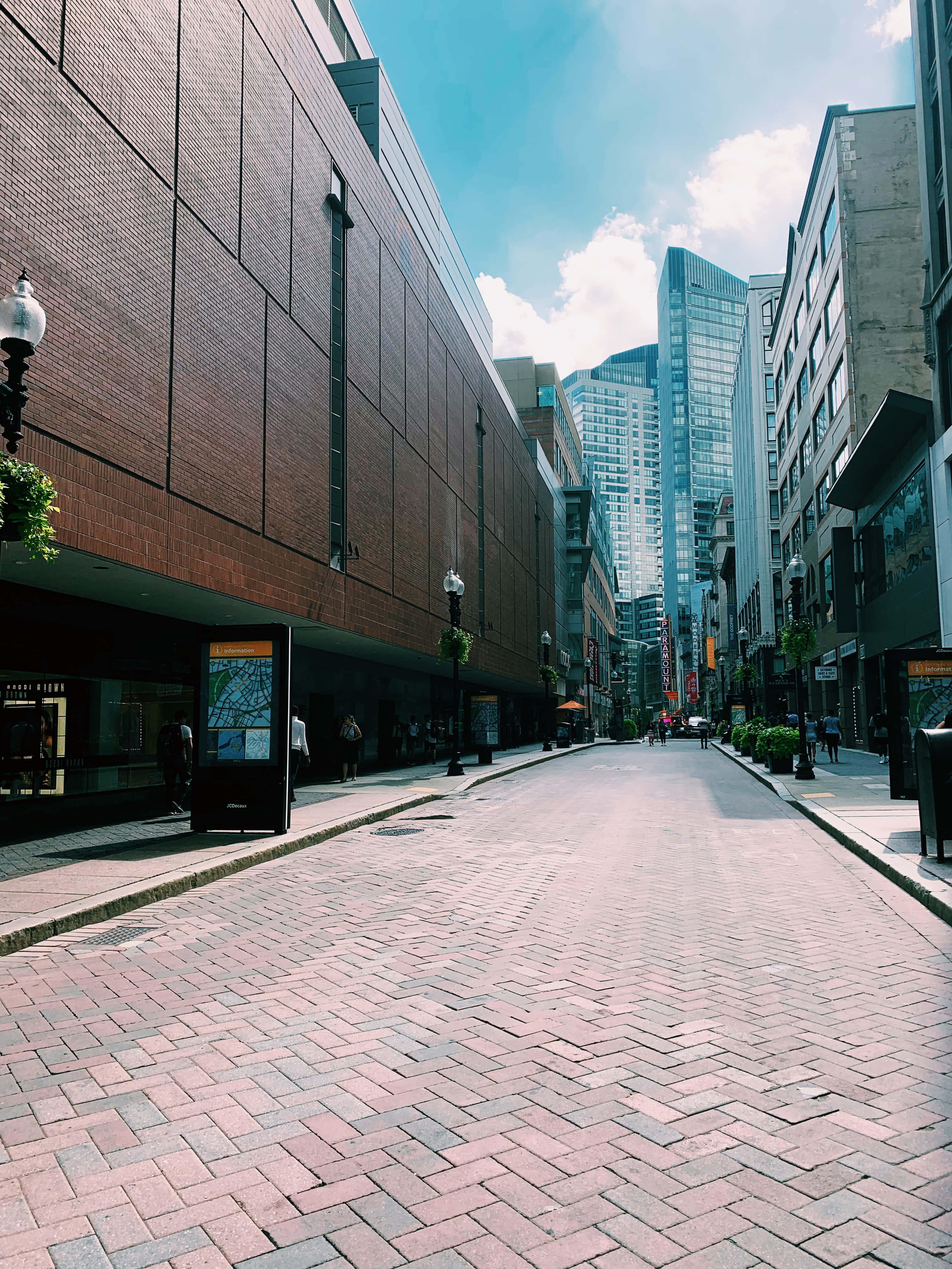 Sidewalk 桌布 壁紙 — 一條陽光普照、鋪滿磚塊的都市街道，兩旁是現代與歷史建築，上方是晴朗藍天，點綴著幾朵白雲。