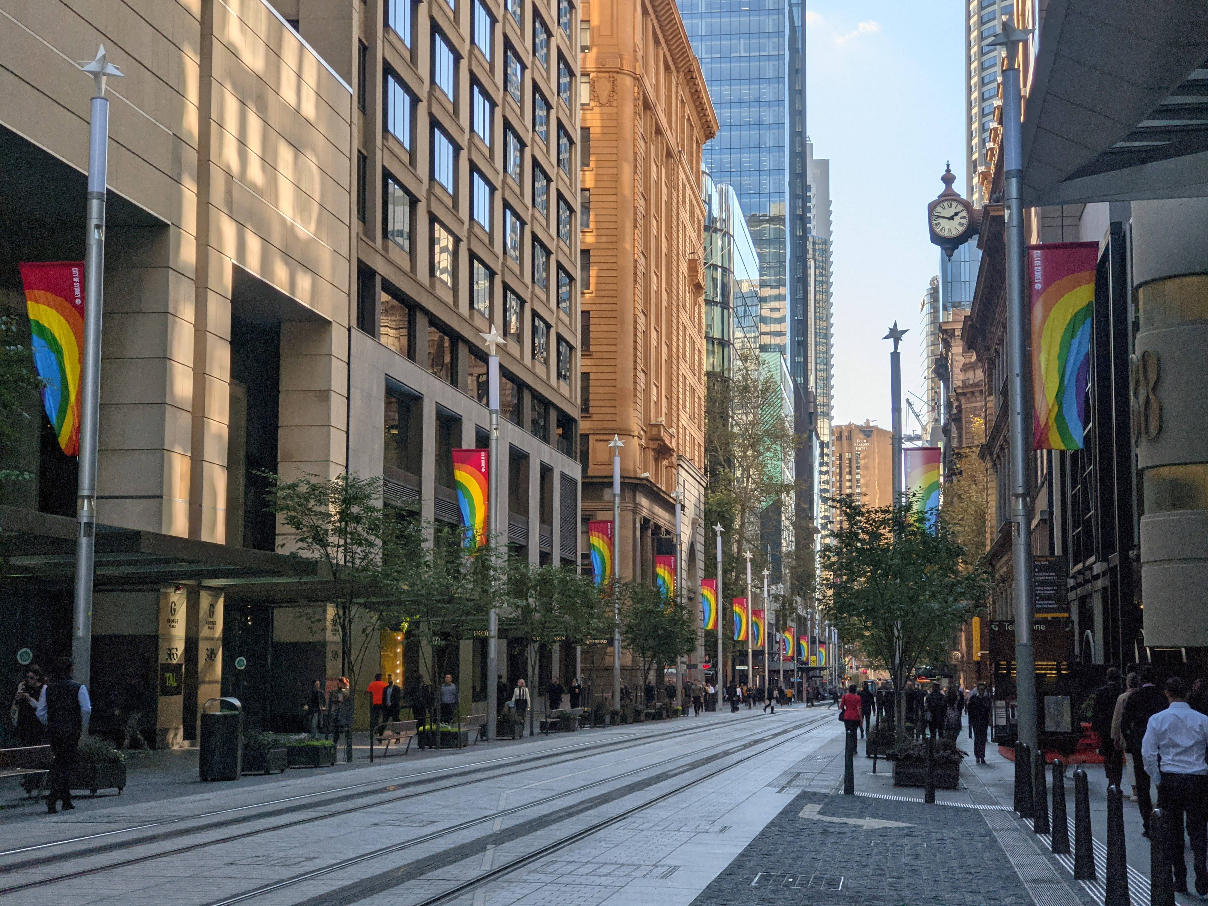 Sydney 桌布 壁紙 — 一條陽光普照的城市街道，兩旁高樓林立，彩虹旗在旗桿上飄揚，行人沿著人行道漫步。
