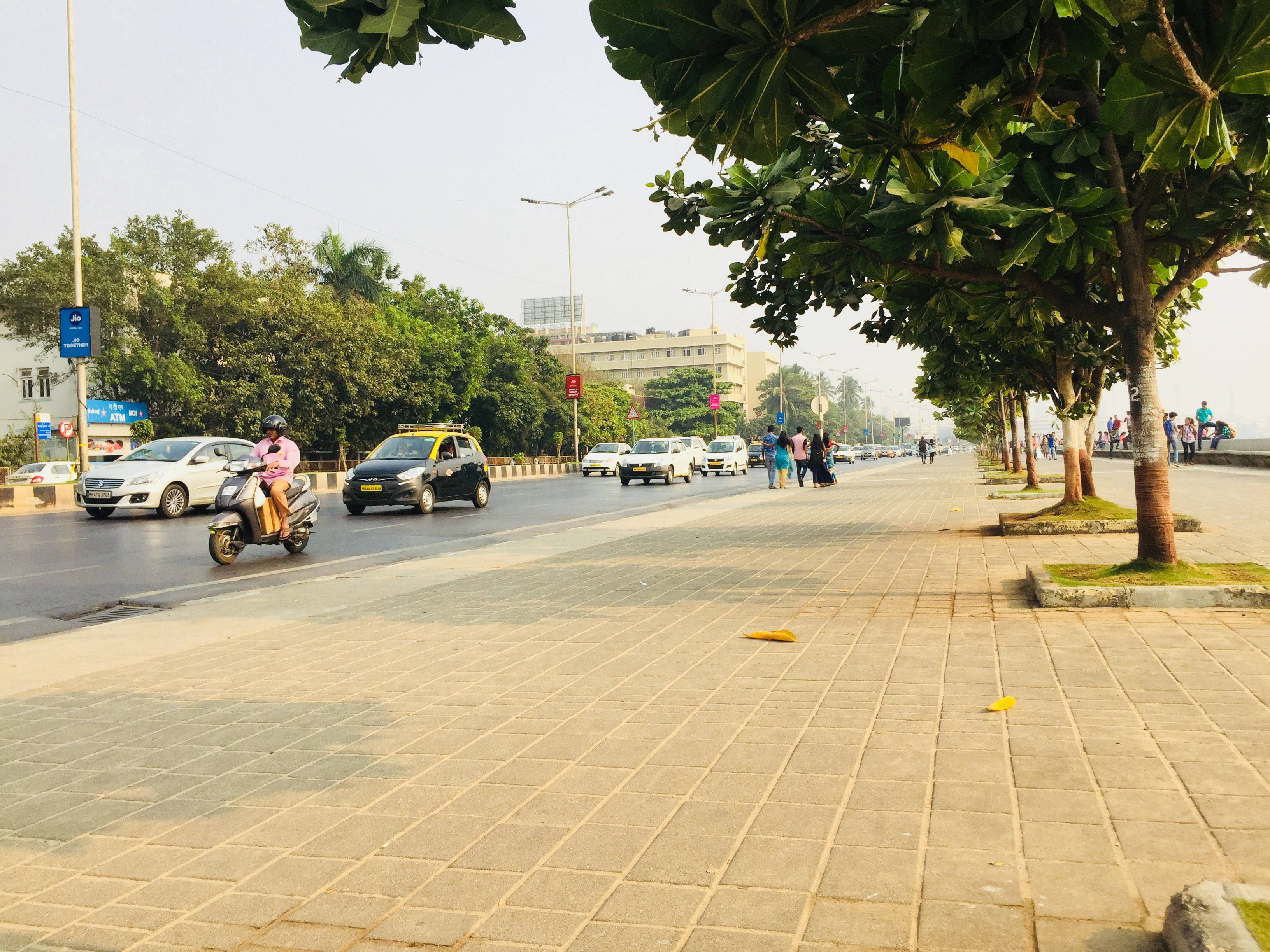 Mumbai 桌布 壁紙 — 一條陽光普照的城市街道，行人與車輛在樹蔭下的人行道上移動，天空略帶朦朧。