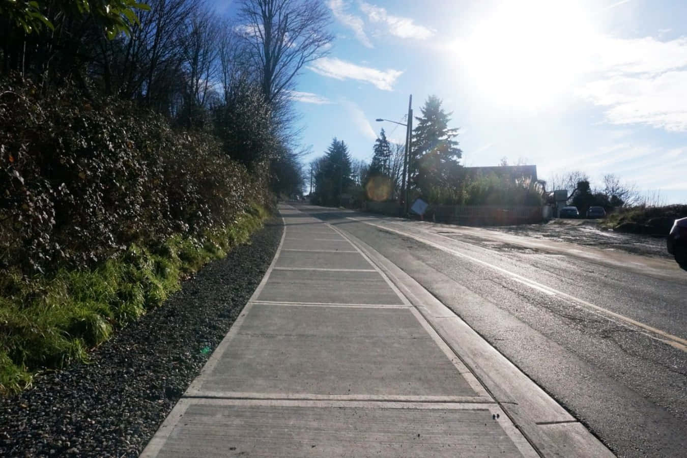 Sidewalk 桌布 壁紙 — 一條陽光普照的柏油道路蜿蜒穿過樹林地區，設有人行道和碎石路肩，上方是明亮的藍天。
