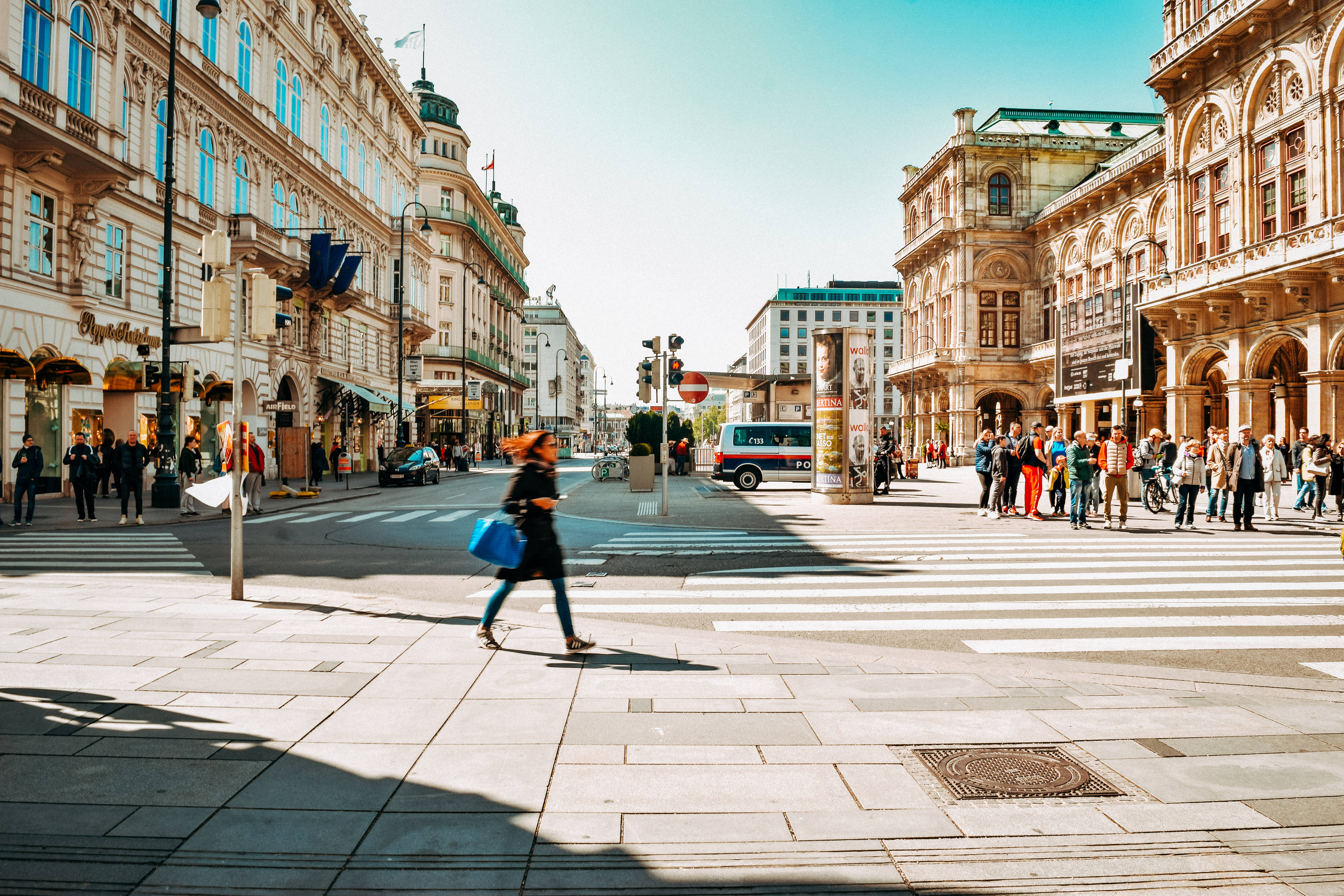 Austria 桌布 壁紙 — 一條陽光普照的歐洲城市街道，有華麗的建築物、行人正在過斑馬線，以及一位正在移動的女性手持一個藍色袋子。
