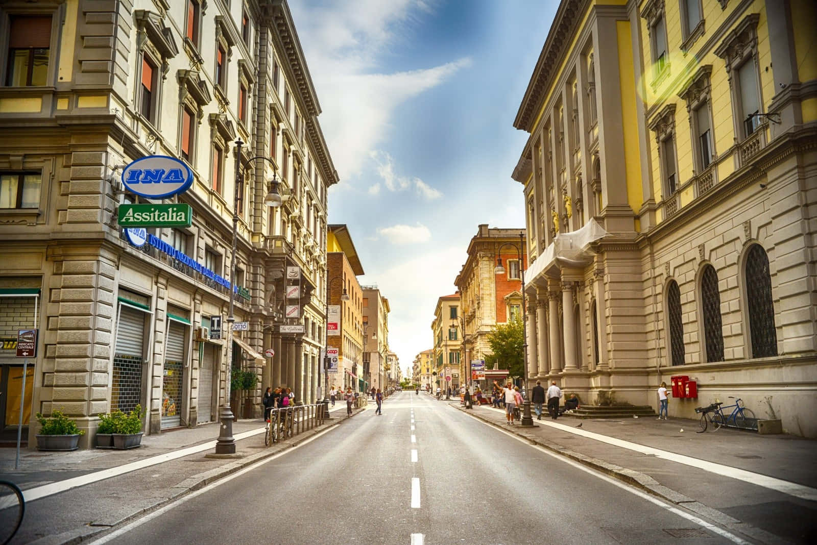 Street 桌布 壁紙 — 一條陽光普照的歐洲街道，兩旁是歷史建築，行人如織，頭頂是湛藍的天空，點綴著幾朵白雲。