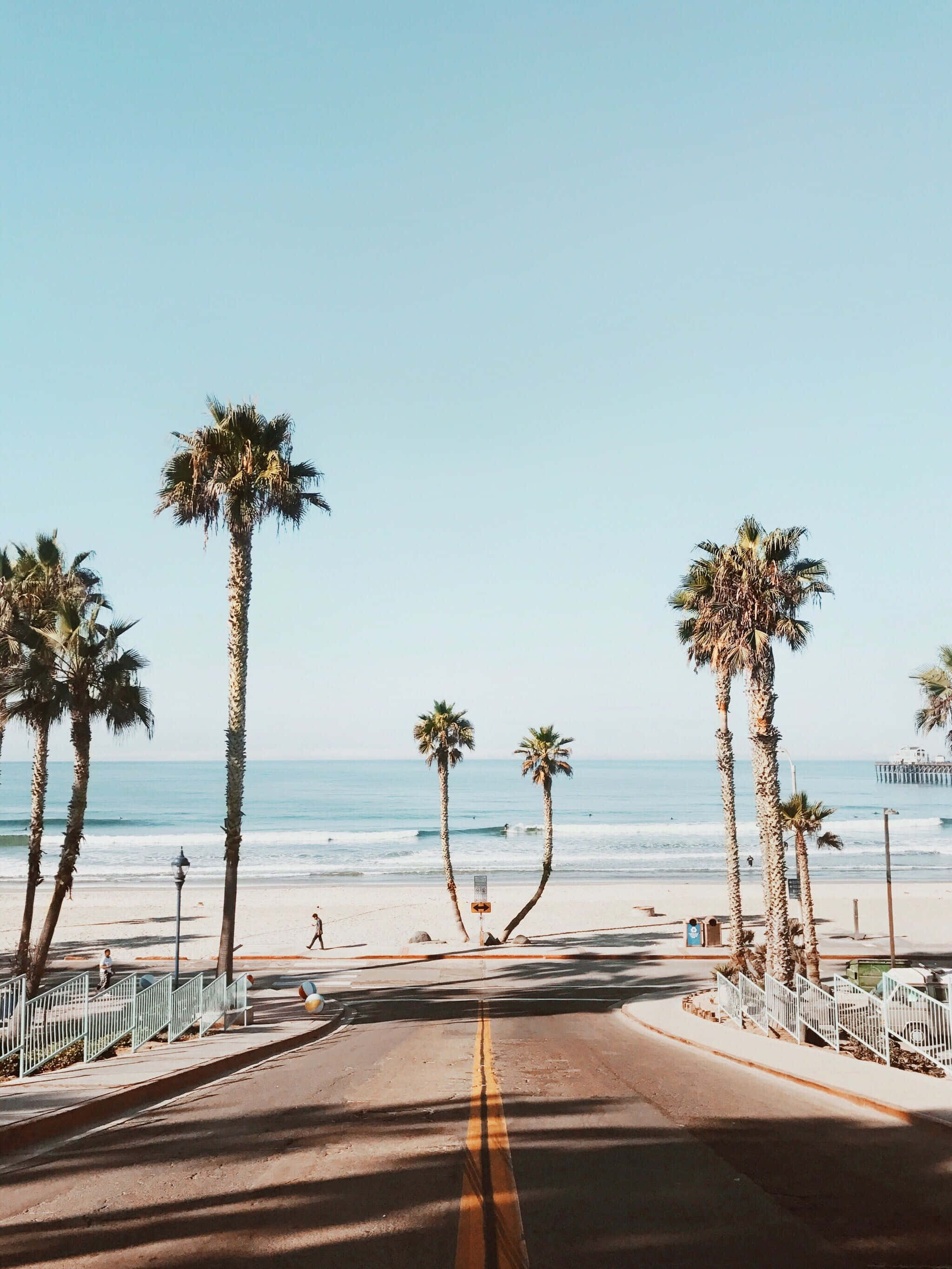 California Aesthetic 桌布 壁紙 — 一條陽光普照的海岸道路，兩旁種滿高大的棕櫚樹，通向沙灘和海洋，天空清澈湛藍。