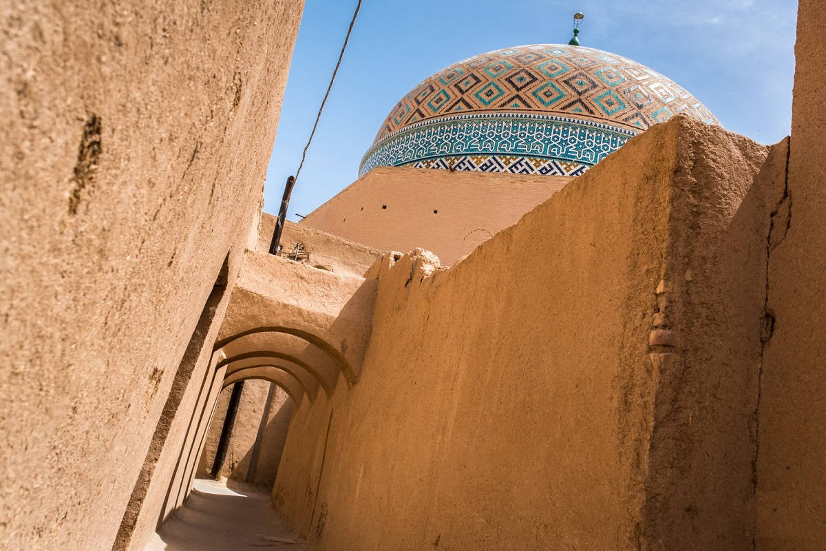 Iran 桌布 壁紙 — 一條陽光普照的狹窄小巷，兩旁是風化的土坯牆，通往晴朗天空下色彩鮮豔的藍金色瓷磚圓頂。