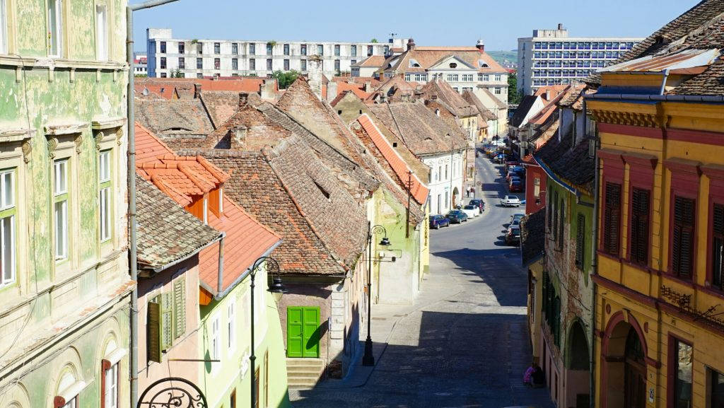 Romania 桌布 壁紙 — 一條陽光普照的狹窄街道蜿蜒於歐洲小鎮中色彩繽紛的歷史建築之間，這些建築擁有赤陶屋頂。