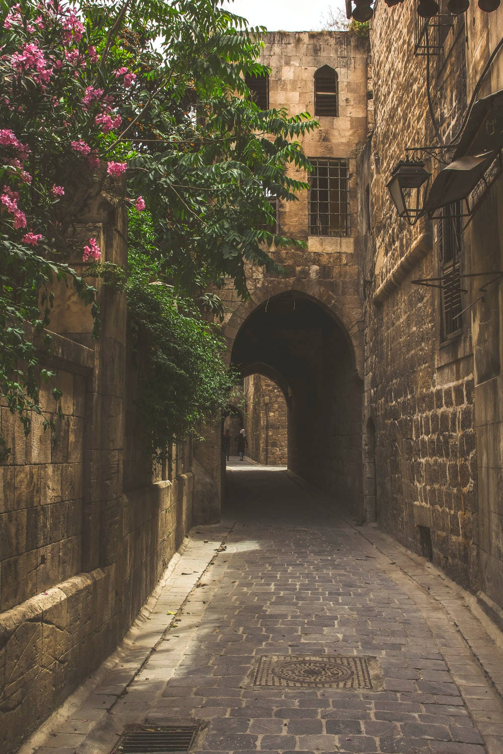 Syria 桌布 壁紙 — 一條陽光普照的鵝卵石小巷，位於古老的石頭城市，兩旁盛開的粉紅色花朵和拱形大門構成框架。
