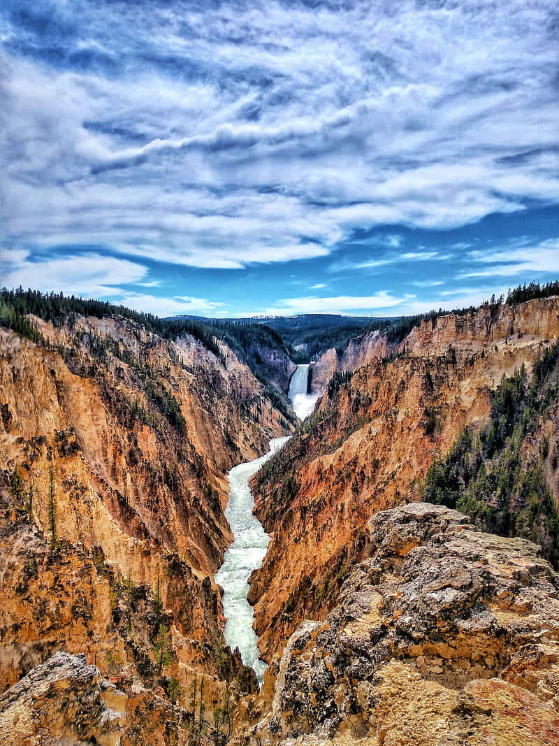 Yellowstone 桌布 壁紙 — 一條雄偉的河流穿過一個深邃的峽谷，兩旁是橙色的懸崖，遠處有瀑布，在戲劇性的藍天下。