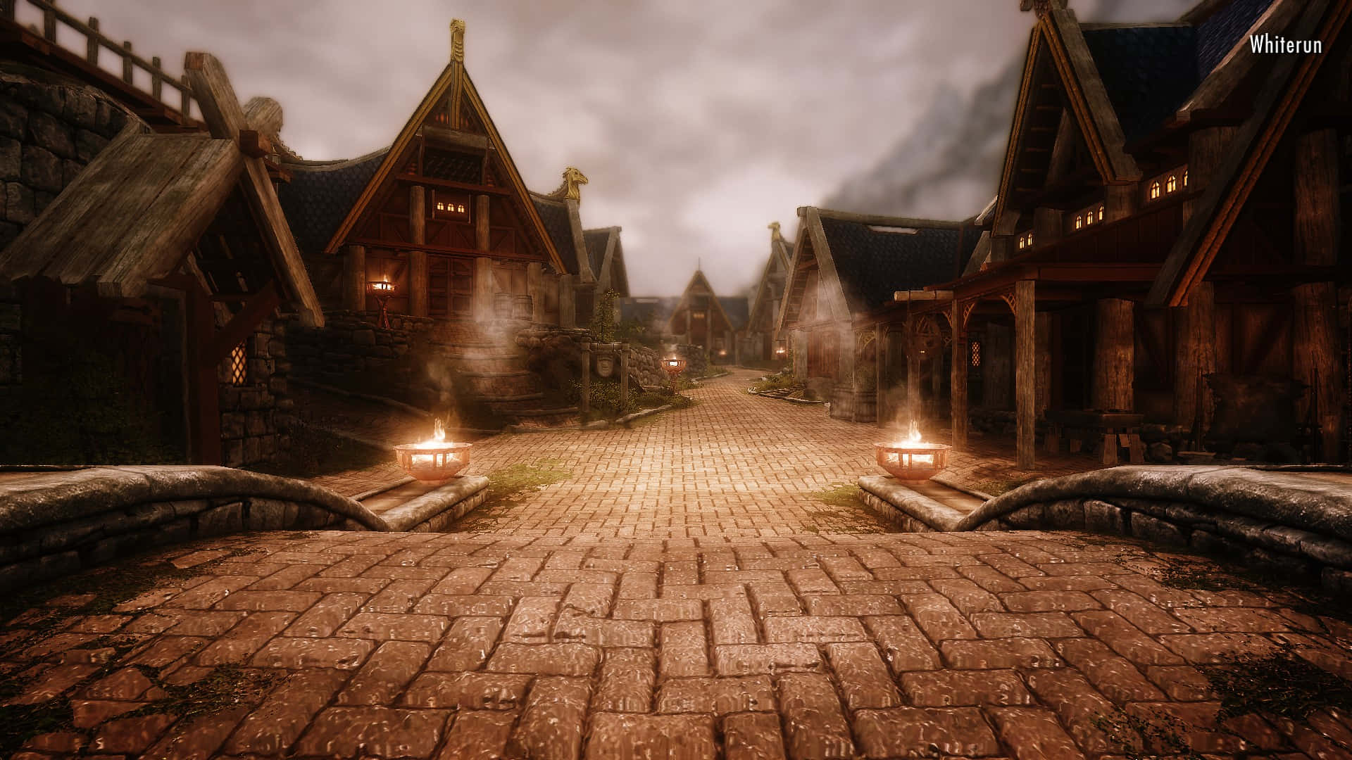 Skyrim Whiterun 桌布 壁紙 — 一條霧氣瀰漫的中世紀村莊街道，兩旁是石頭小屋和發光的燈籠，天空陰沉。