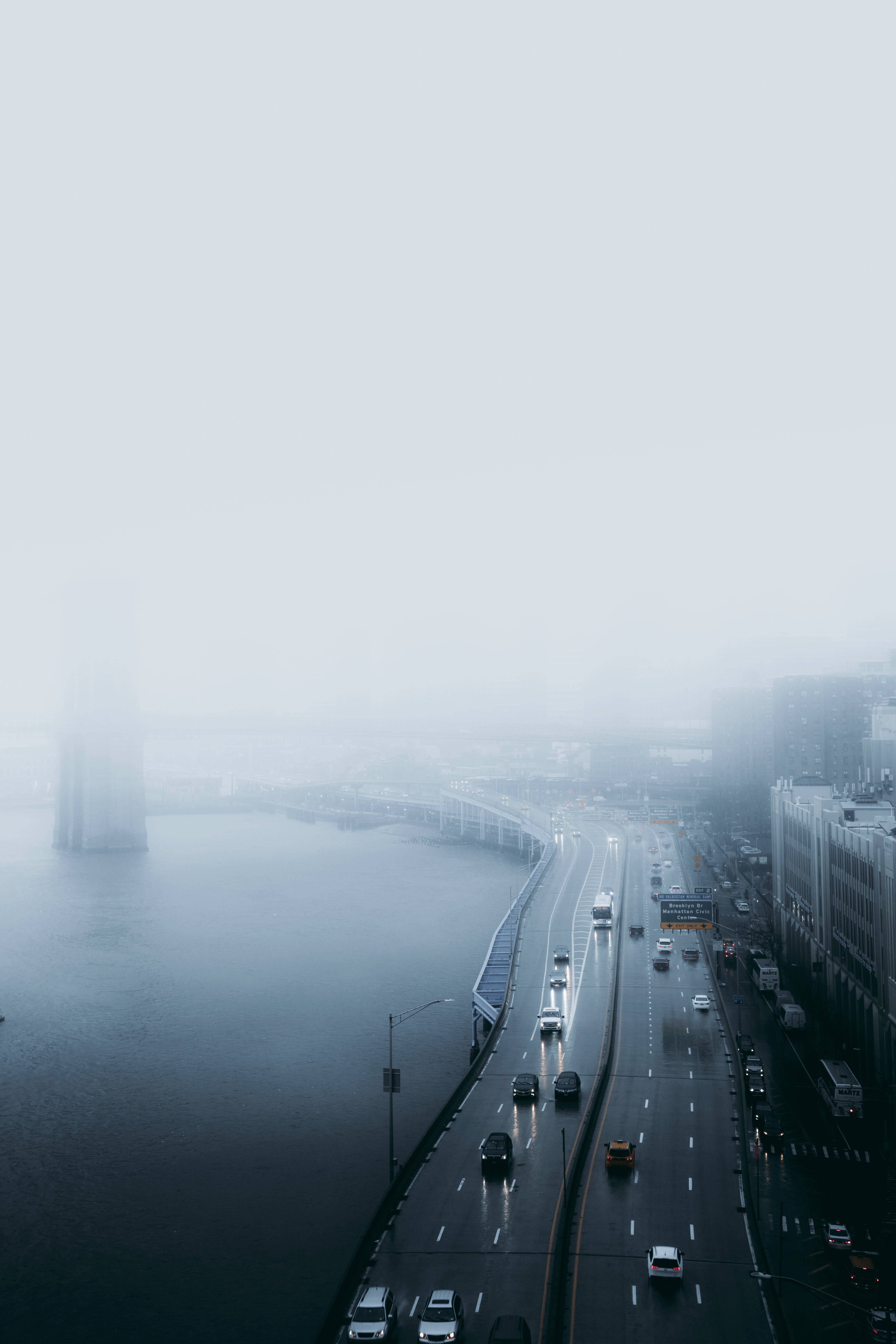 Raining 桌布 壁紙 — 一條霧氣瀰漫的城市高速公路沿著平靜的水道蜿蜒而行，透過霧氣隱約可見城市的建築物。