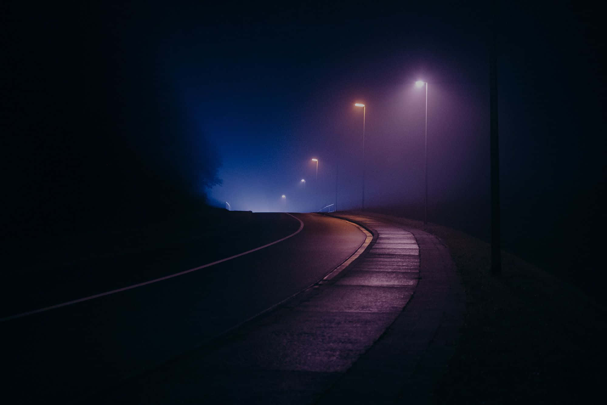 Romanticism 桌布 壁紙 — 一條霧氣瀰漫的夜路在發光的路燈下輕輕彎曲，透過濃霧投射出紫色和藍色的色調。