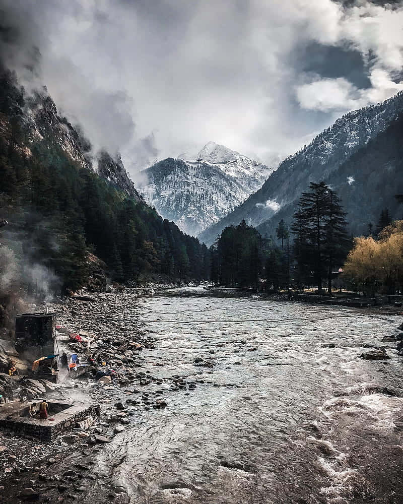 Himalaya 桌布 壁紙 — 一條霧氣瀰漫的山間河流流經岩石山谷，兩旁有積雪的山峰和常綠樹木，天空多雲。