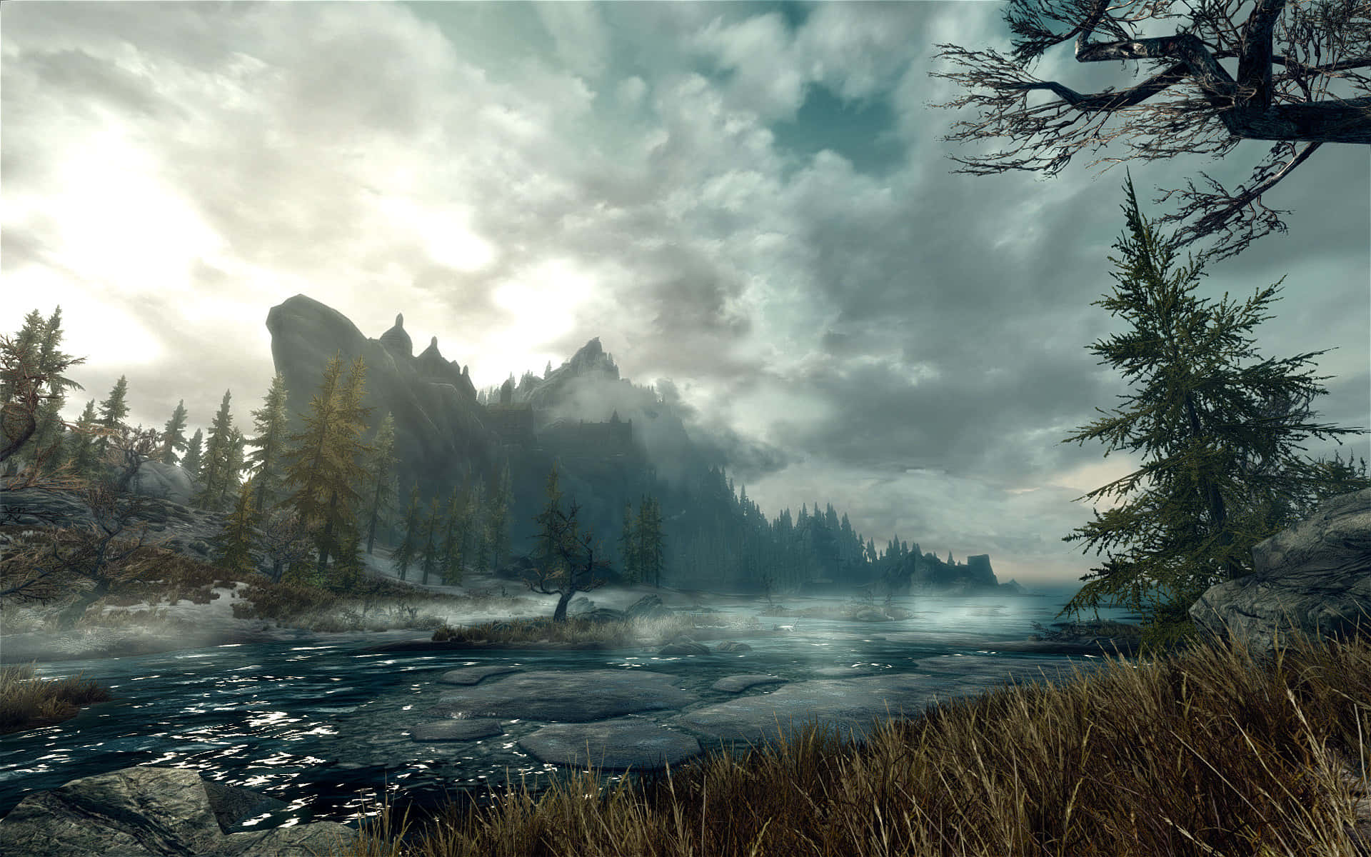 Skyrim Landscapes 桌布 壁紙 — 一條霧氣瀰漫的河流蜿蜒穿過白雪覆蓋的森林，高聳的山脈在陰雲密布的天空下。