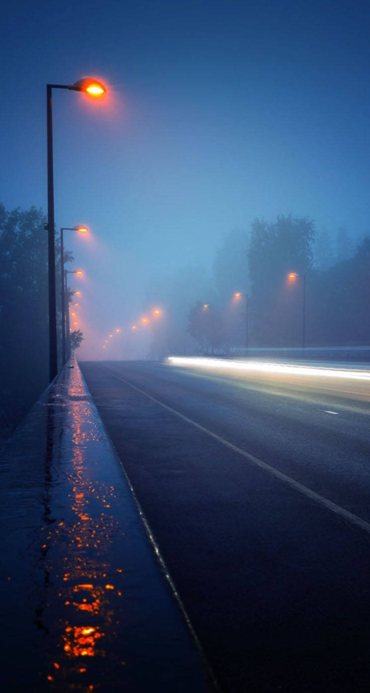 桌布 壁紙 — 一條霧氣瀰漫的黃昏道路，被發光的街燈照亮，倒映在濕漉漉的路面上，並有一道汽車燈光軌跡。