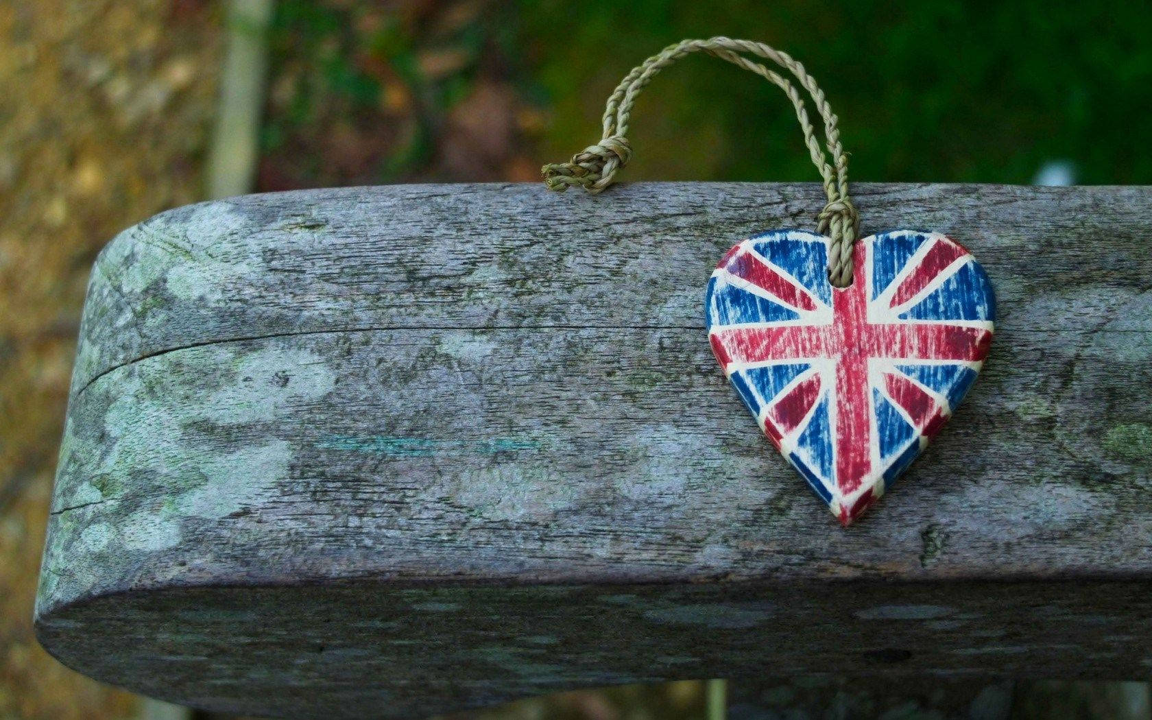United Kingdom Flag 桌布 壁紙 — 一條風化的木樑上，用麻繩繫著一個繪有英國國旗的心形裝飾，背景是模糊的自然景色。