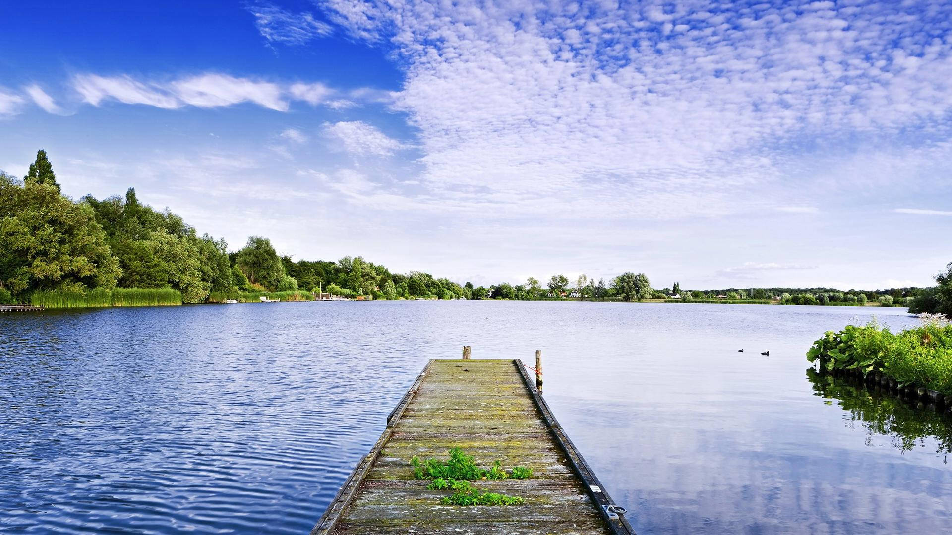 Lake 桌布 壁紙 — 一條風化的木製碼頭延伸到平靜的藍色湖泊，天空明亮，散佈著白雲。