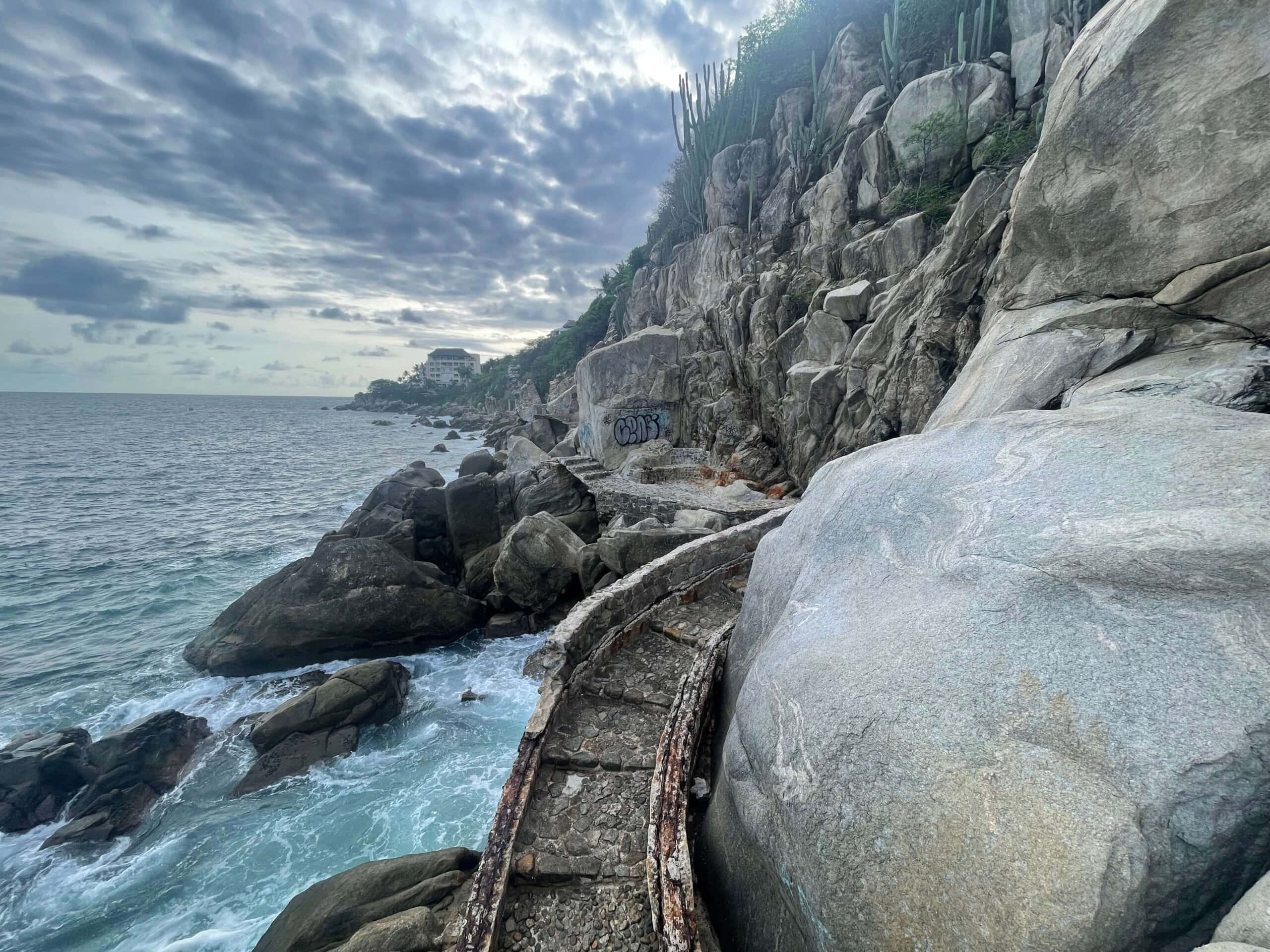 Seaside 桌布 壁紙 — 一條風化的石徑蜿蜒於崎嶇的懸崖邊，在陰天之下，碧綠色的海浪拍打著灰色的巨石。