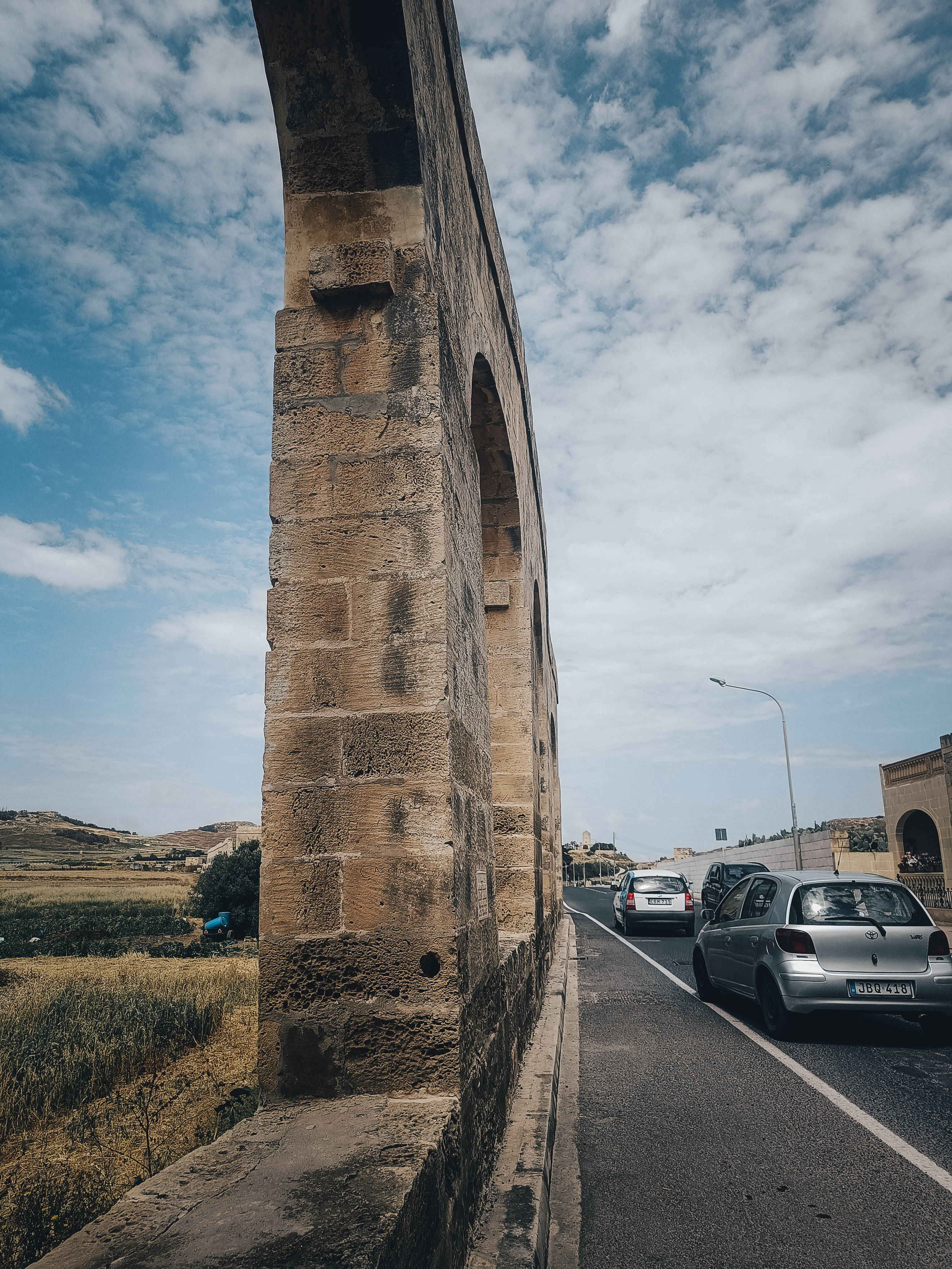 Malta 桌布 壁紙 — 一條風化的石造高架渠拱起在道路上，汽車在部分多雲的藍天下駛過。