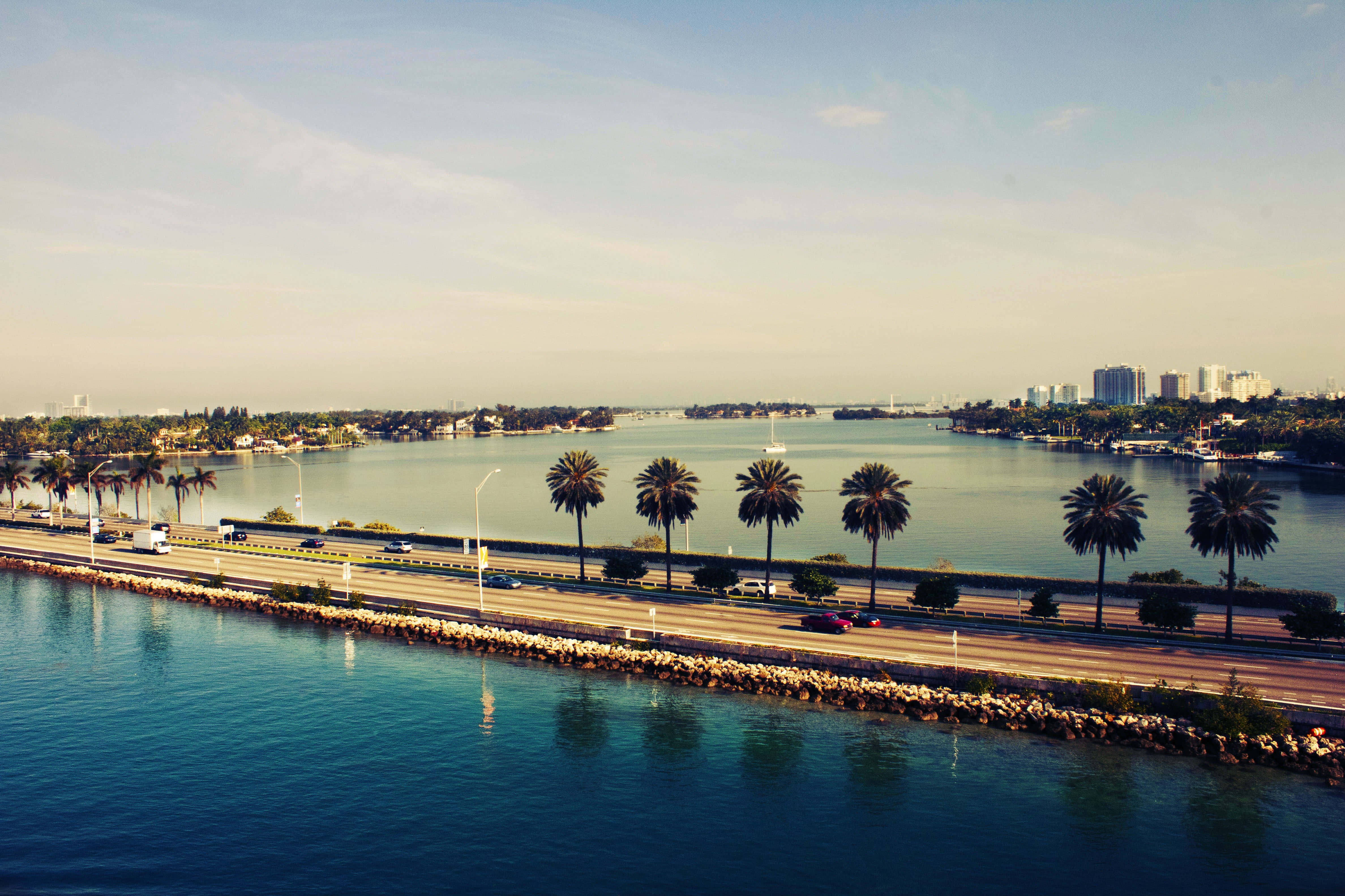 Miami 桌布 壁紙 — 一條風景優美的沿海公路兩旁種滿棕櫚樹，沿著平靜的藍色海水延伸，遠處的城市建築在晴朗的天空下清晰可見。