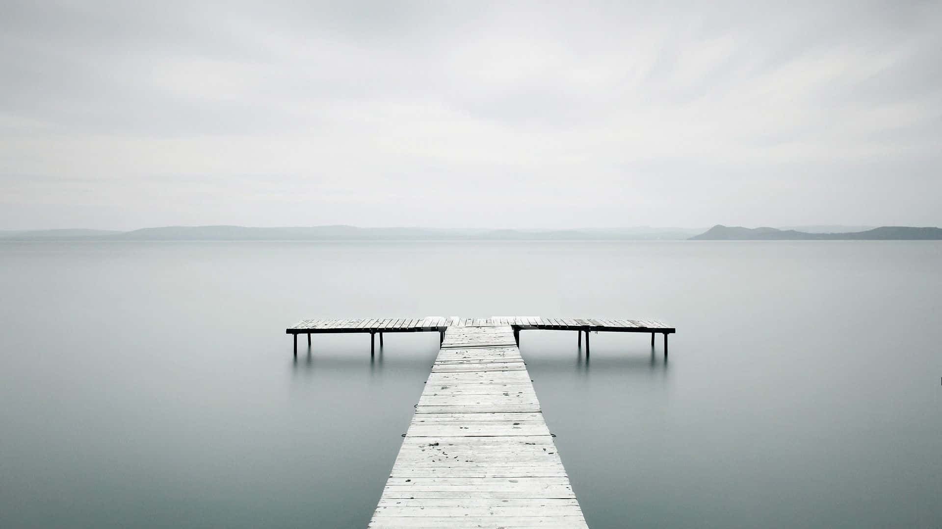 Minimalism 桌布 壁紙 — 一條飽經風霜的木碼頭延伸到平靜的灰色湖面上，天空多雲，地平線上有遠山。