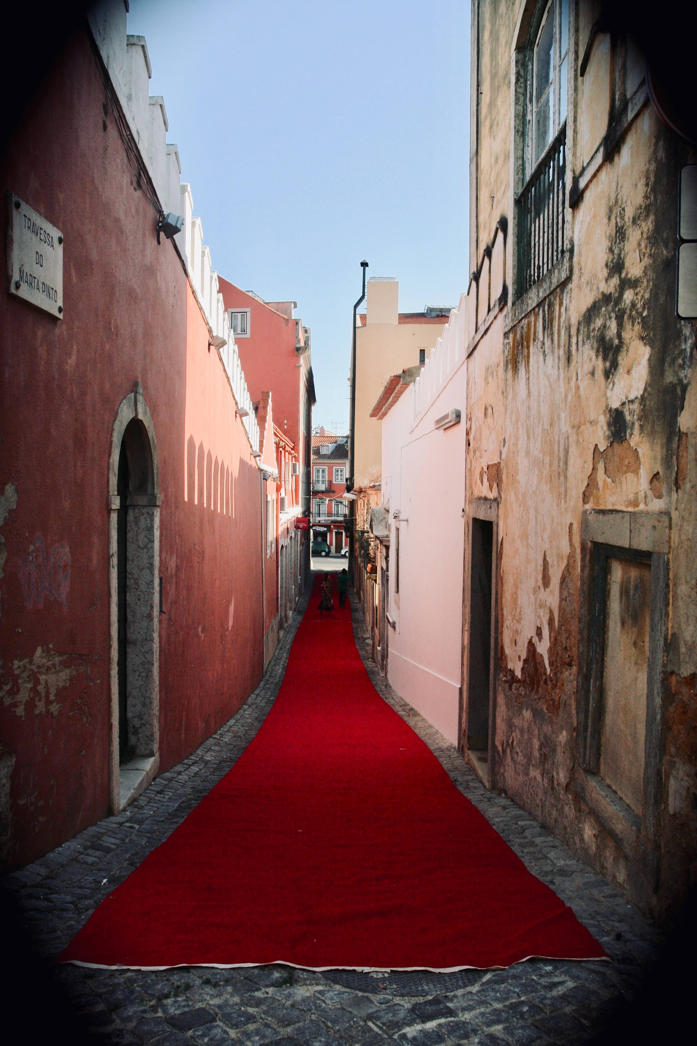 Red Carpet 桌布 壁紙 — 一條鮮紅色的地毯在晴朗的藍天下，沿著一條狹窄的鵝卵石小巷，在風化的粉紅色和赭色建築物之間延伸。