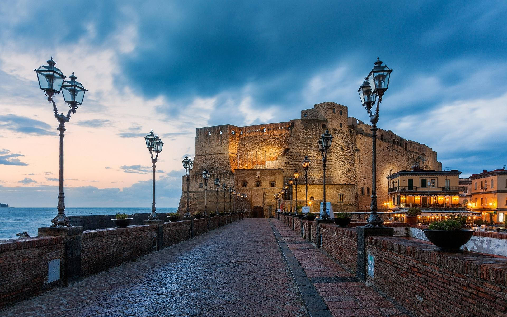 Naples 桌布 壁紙 — 一條鵝卵石小徑在暮色中通往一座宏偉的石堡，溫暖的燈光在戲劇性的藍天下閃爍。