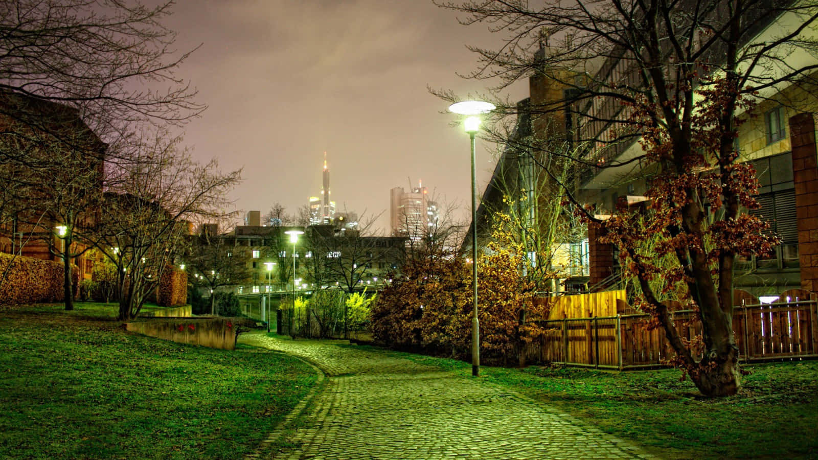Fall City 桌布 壁紙 — 一條鵝卵石小徑蜿蜒穿過夜晚薄霧籠罩的秋日公園，光禿的樹木和發光的街燈勾勒出遠處的城市天際線。