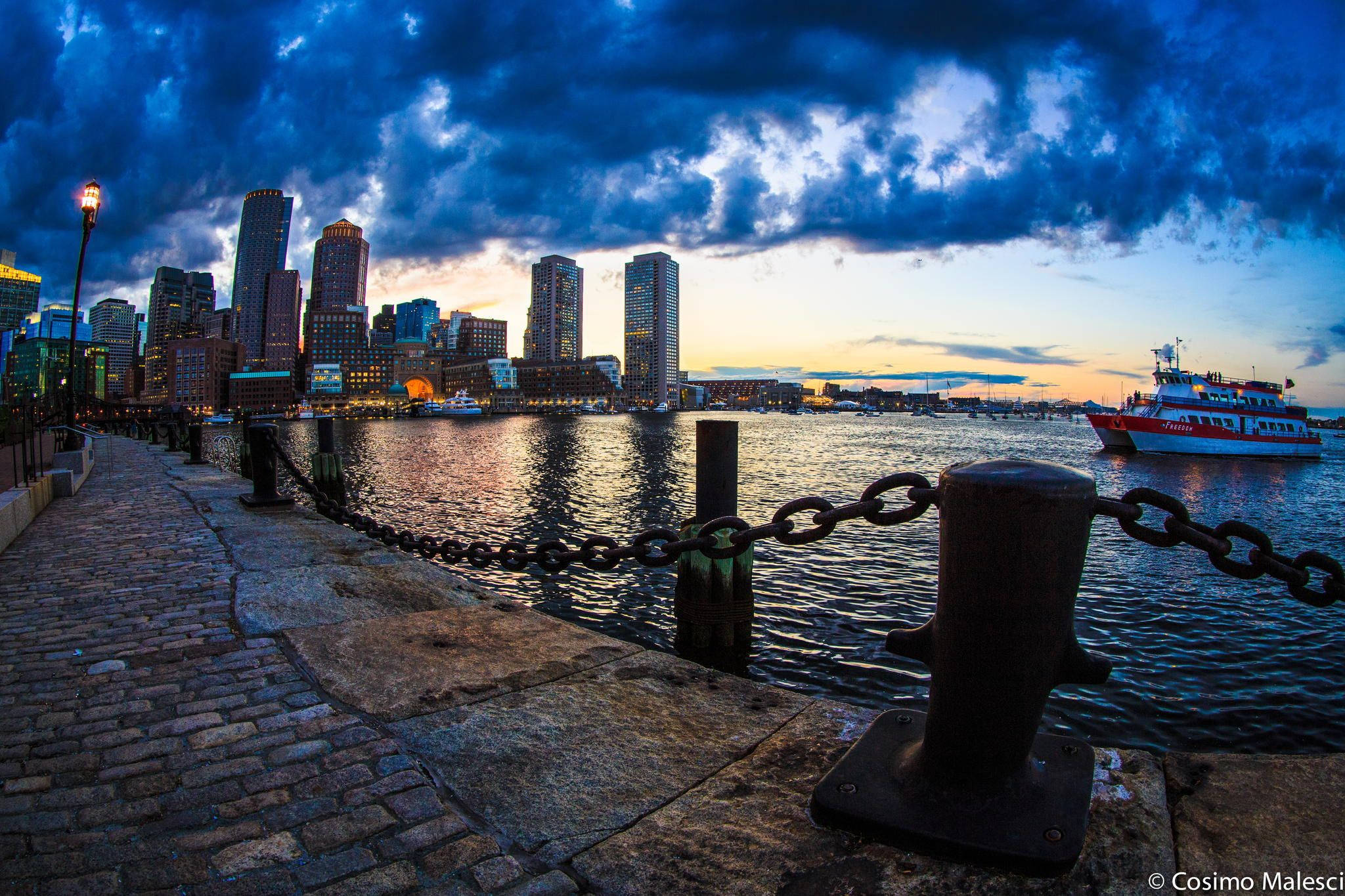 Boston 桌布 壁紙 — 一條鵝卵石海濱小徑通向黃昏時分的城市天際線，天空中有戲劇性的雲彩，水面上有一艘紅色渡輪。