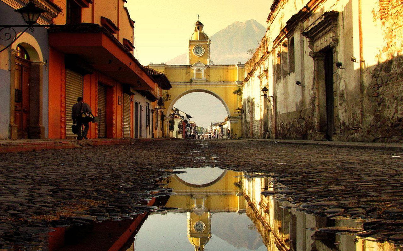 Guatemala 桌布 壁紙 — 一條鵝卵石街道在金色夕陽下映照出歷史悠久的黃色拱門和遠山。