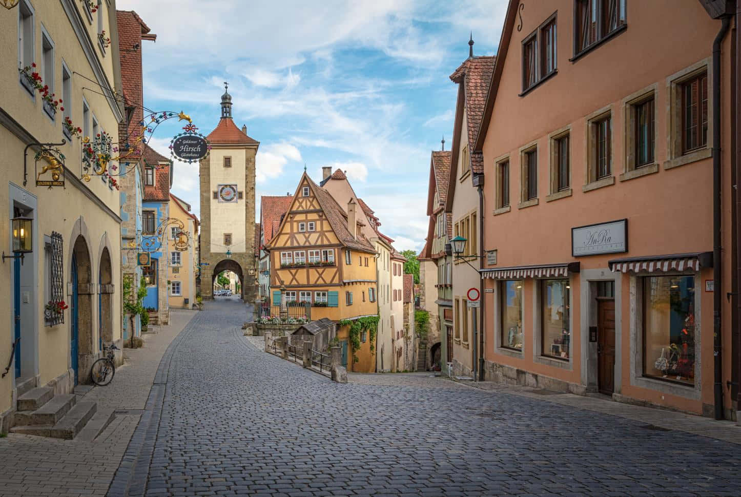German 桌布 壁紙 — 一條鵝卵石街道蜿蜒穿過色彩繽紛的半木結構房屋，通往一座歷史悠久的石塔，上方是明亮的藍天。