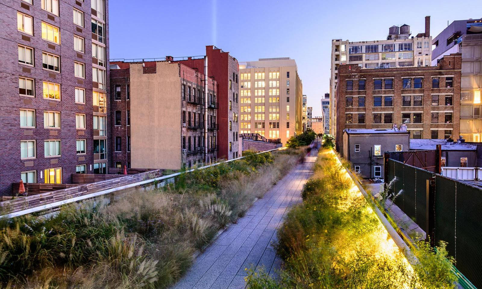 New York 桌布 壁紙 — 一條黃昏時分的高架公園小徑蜿蜒於城市建築之間，建築物窗戶閃爍著光芒，小徑上有金色的燈光。