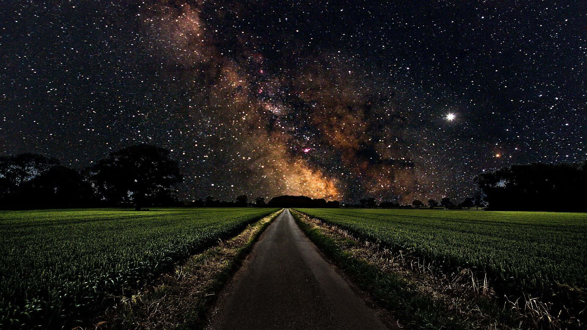 星空 桌布 壁紙 — 一條黑暗蜿蜒的道路穿過綠色的田野，上方是令人驚嘆的星空，銀河在頭頂閃耀。