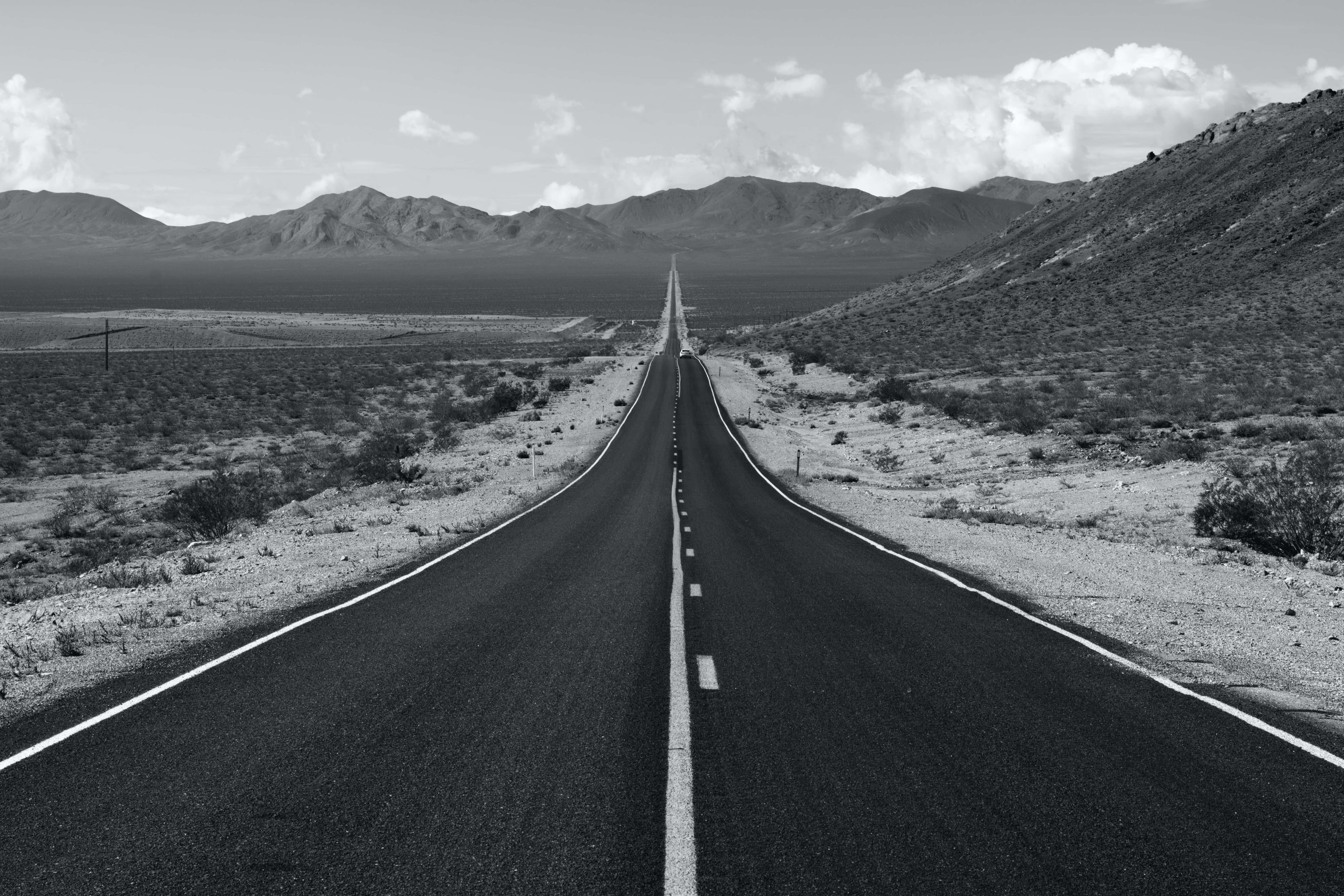 Aesthetic Black And White 桌布 壁紙 — 一條黑白相間的筆直道路，在陰天之下，無盡地延伸穿過荒蕪的沙漠景觀，通往遠方的山脈。