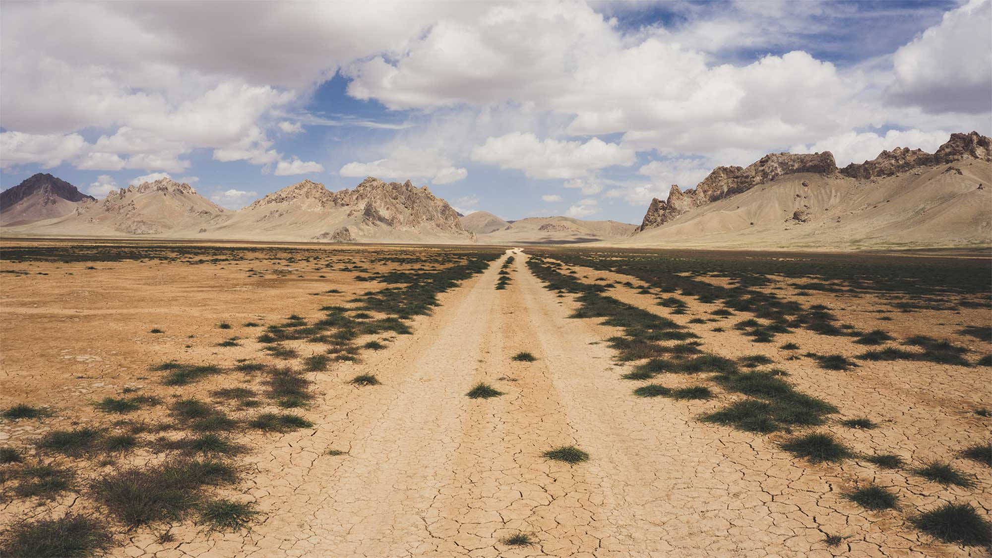 Afghanistan 桌布 壁紙 — 一條龜裂的泥土路在部分多雲的天空下延伸向遠處崎嶇的山脈。