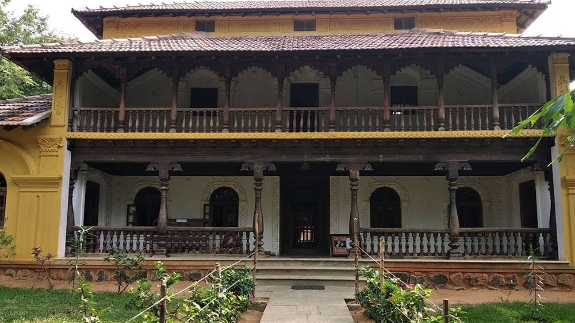 Chennai 桌布 壁紙 — 一棟傳統黃色兩層房屋，設有木製陽台和瓦片屋頂，四周環繞著綠植。