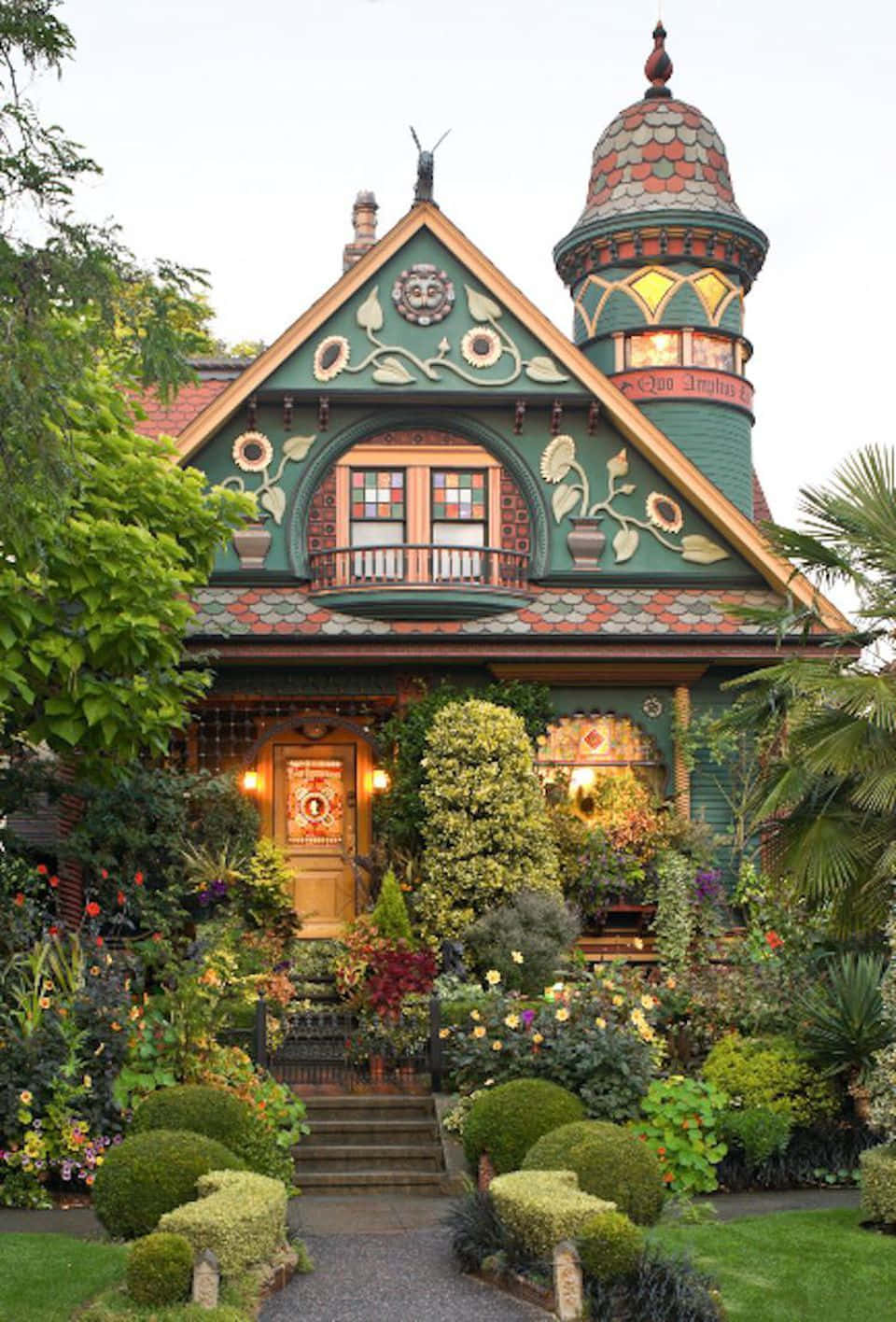 Cottage 桌布 壁紙 — 一棟充滿活力的綠色維多利亞式房屋，帶有華麗的向日葵圖案和瓷磚塔樓，矗立在柔和日光下的茂密花園中。