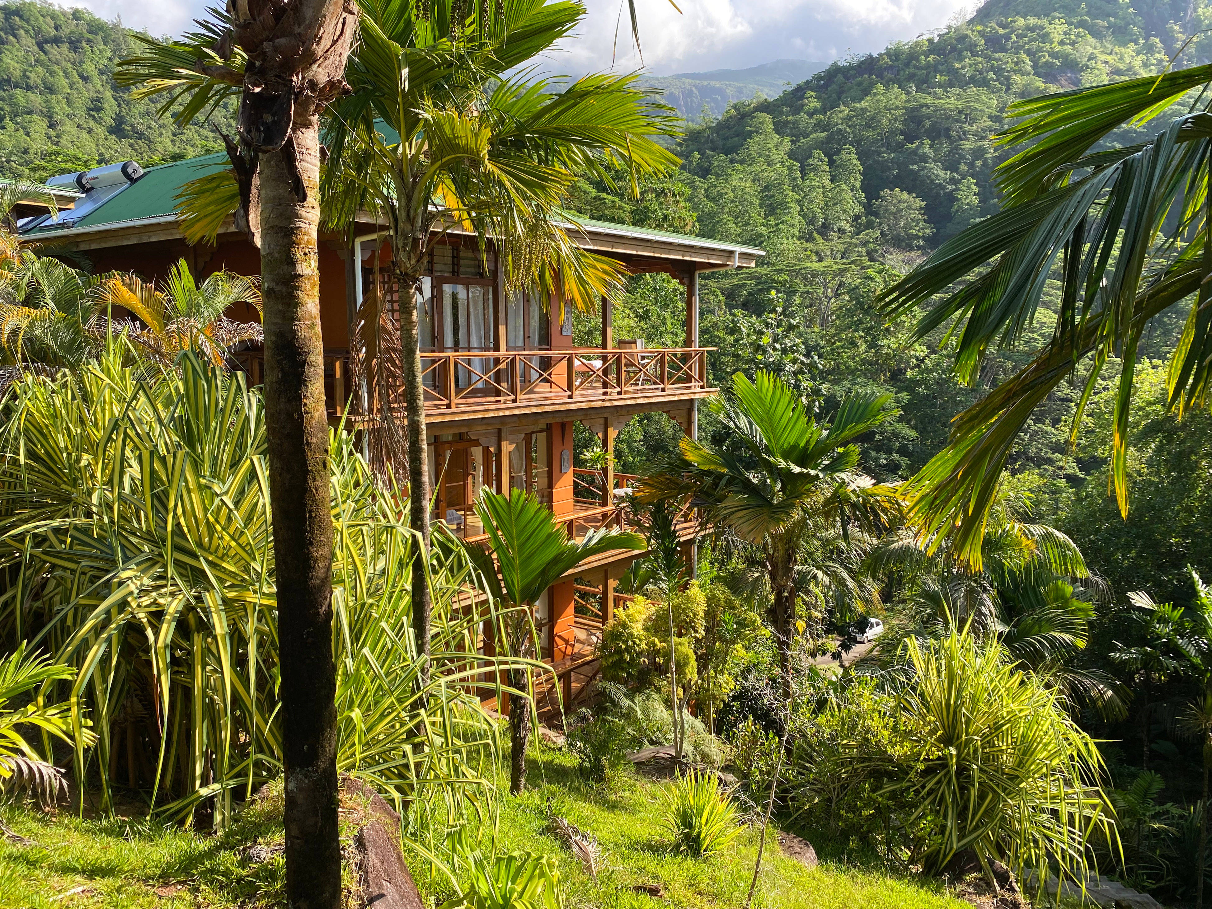 Seychelles 桌布 壁紙 — 一棟兩層樓的木屋，綠色屋頂，坐落在茂密的熱帶棕櫚樹和濃密的綠色山脈之間，沐浴在明亮的日光下。
