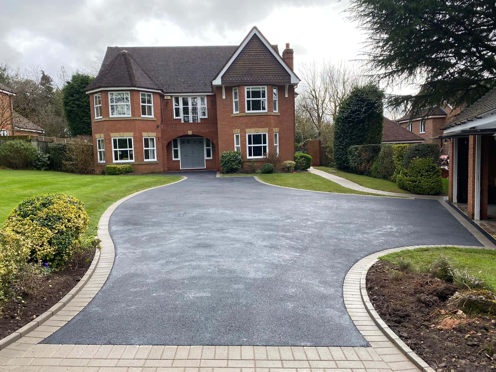 Driveway Ideas 桌布 壁紙 — 一棟帶白色窗戶的大型磚房坐落在新鋪設的彎曲車道盡頭，車道兩旁是修剪整齊的草坪和灌木，天空多雲。
