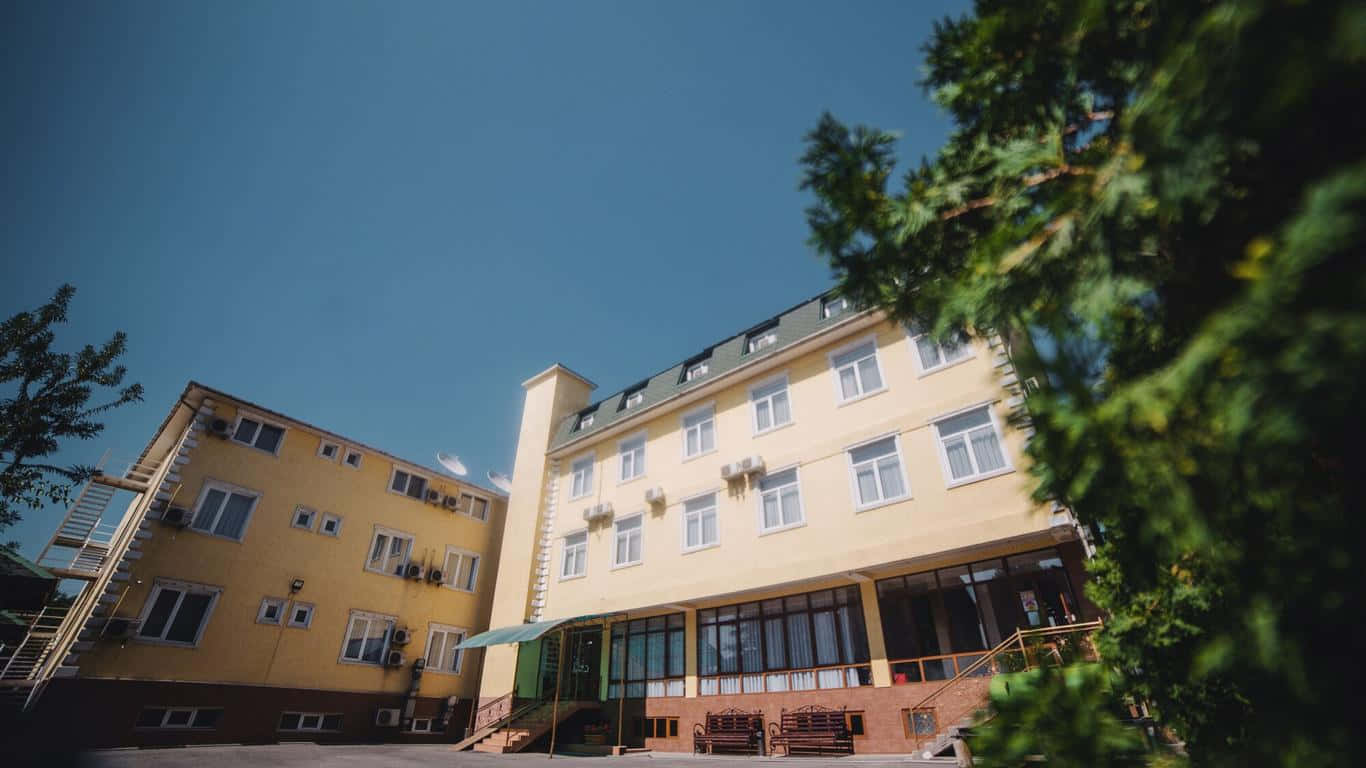 Hotel 桌布 壁紙 — 一棟明亮的黃色多層建築，帶有白色窗戶，矗立在清澈的藍天下，周圍環繞著茂密的綠樹。