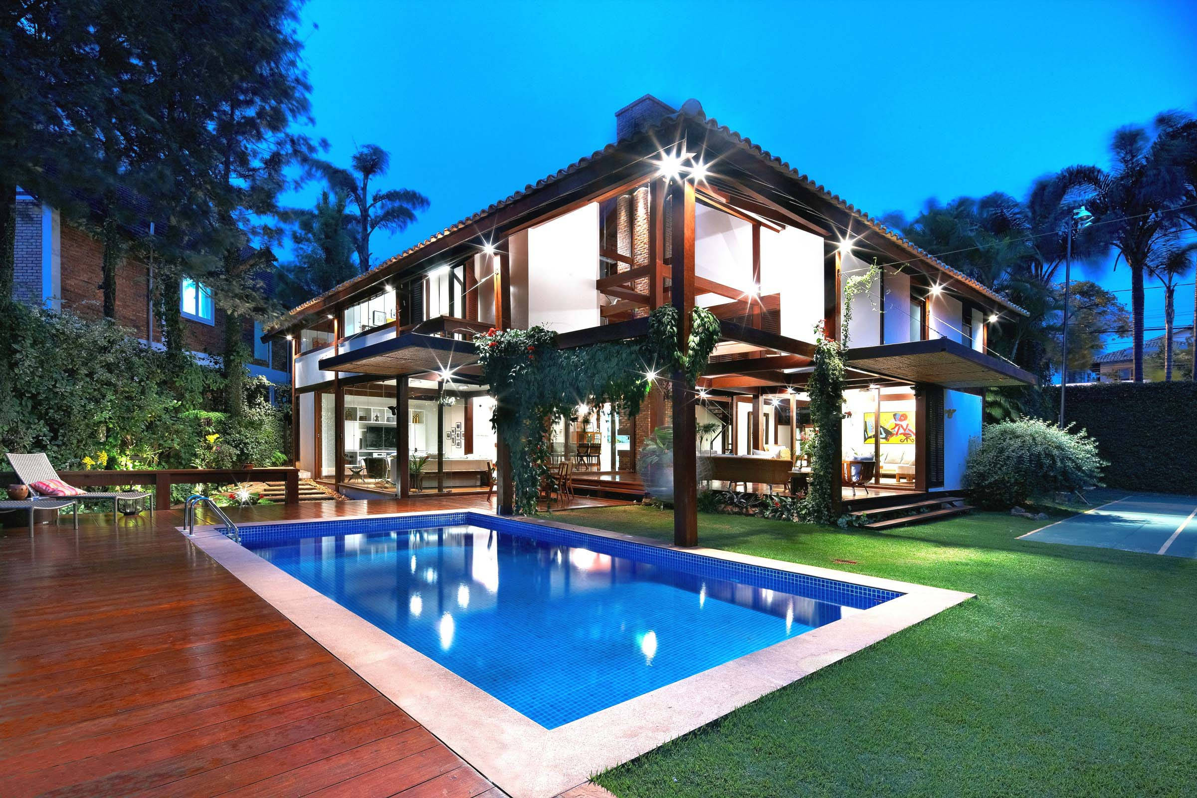 Luxury House 桌布 壁紙 — 一棟現代兩層住宅在黃昏時分亮起溫暖燈光，旁邊是一個充滿活力的藍色泳池，四周環繞著茂密的綠植和木質甲板。