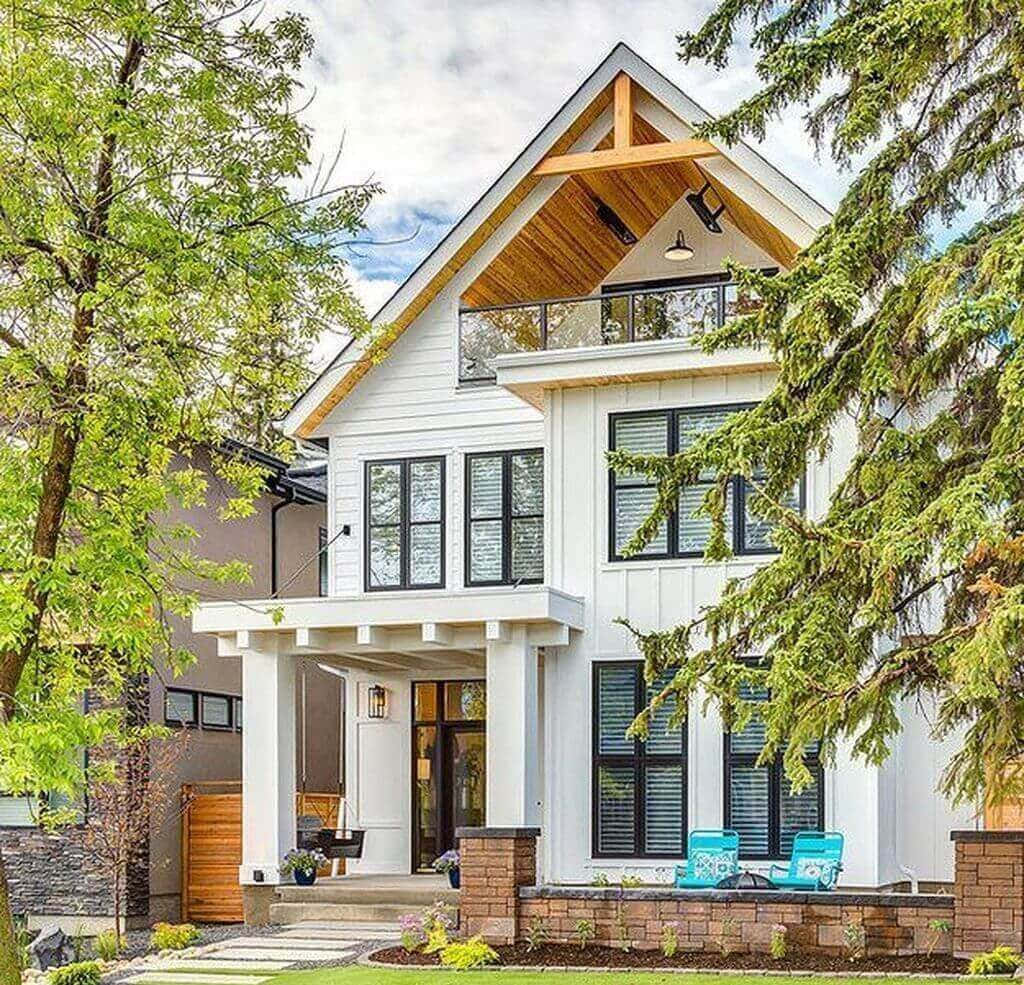 Modern Farmhouse 桌布 壁紙 — 一棟現代白色房屋，配有黑色窗戶和木質裝飾，坐落在茂密的綠樹之中，天空部分多雲。