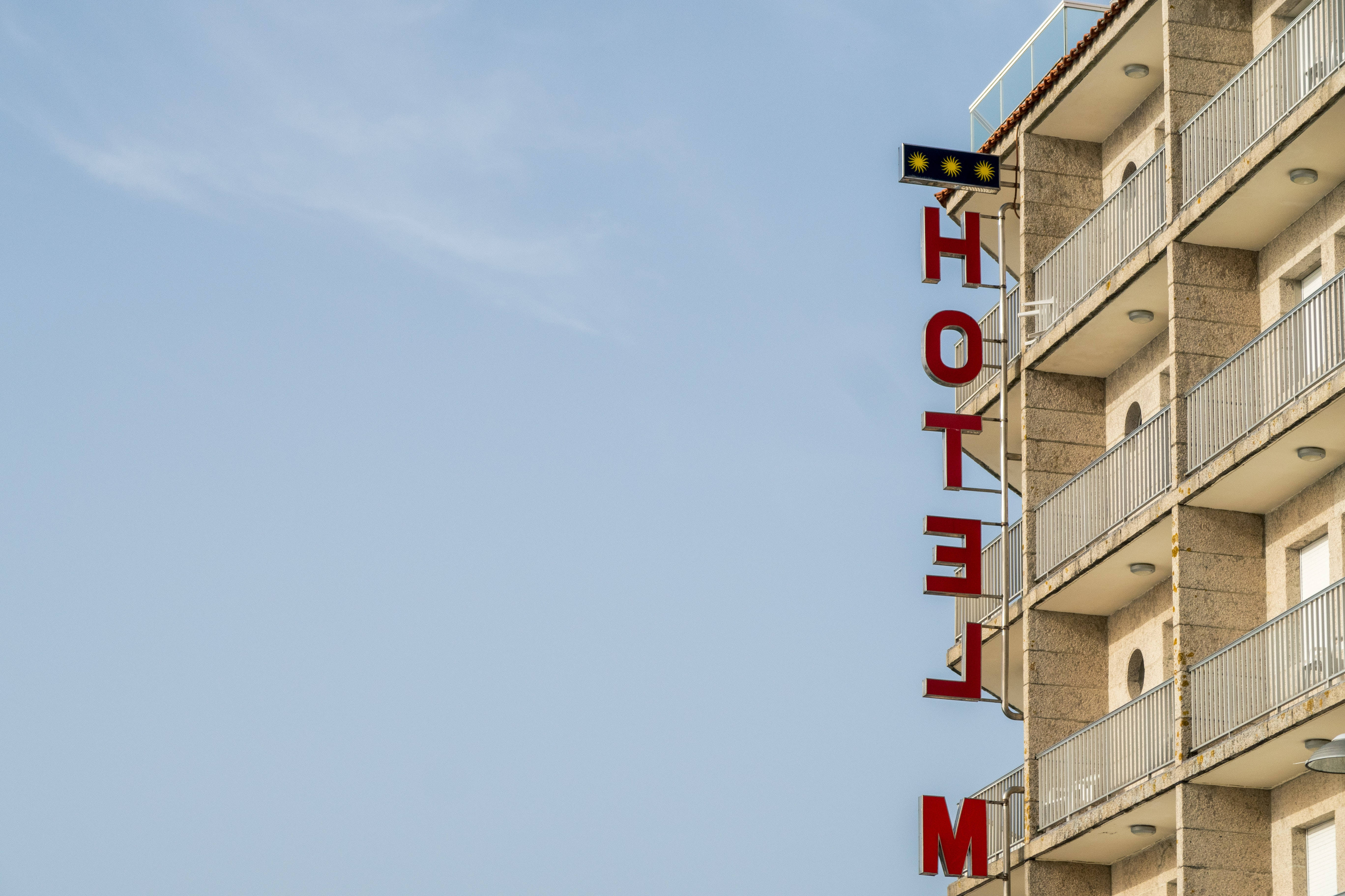 Hotel 桌布 壁紙 — 一棟米色高層酒店建築，在晴朗的藍天下豎立著一個紅色的垂直「HOTEL」標誌。