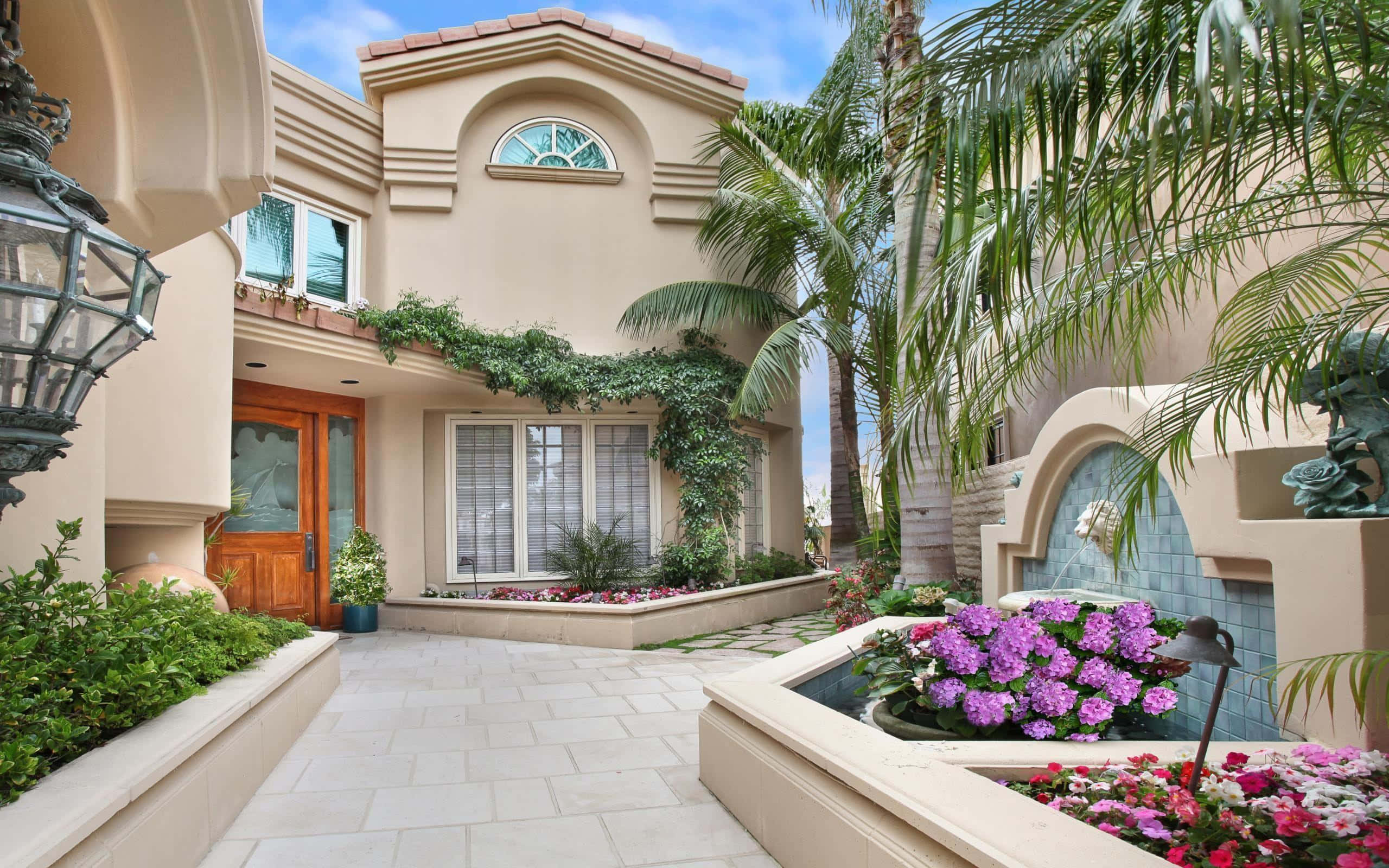 Beautiful House 桌布 壁紙 — 一棟豪華的米色地中海風格住宅，擁有拱形窗戶和瓷磚庭院，周圍環繞著棕櫚樹和充滿活力的紫色花朵，在清澈的藍天下。