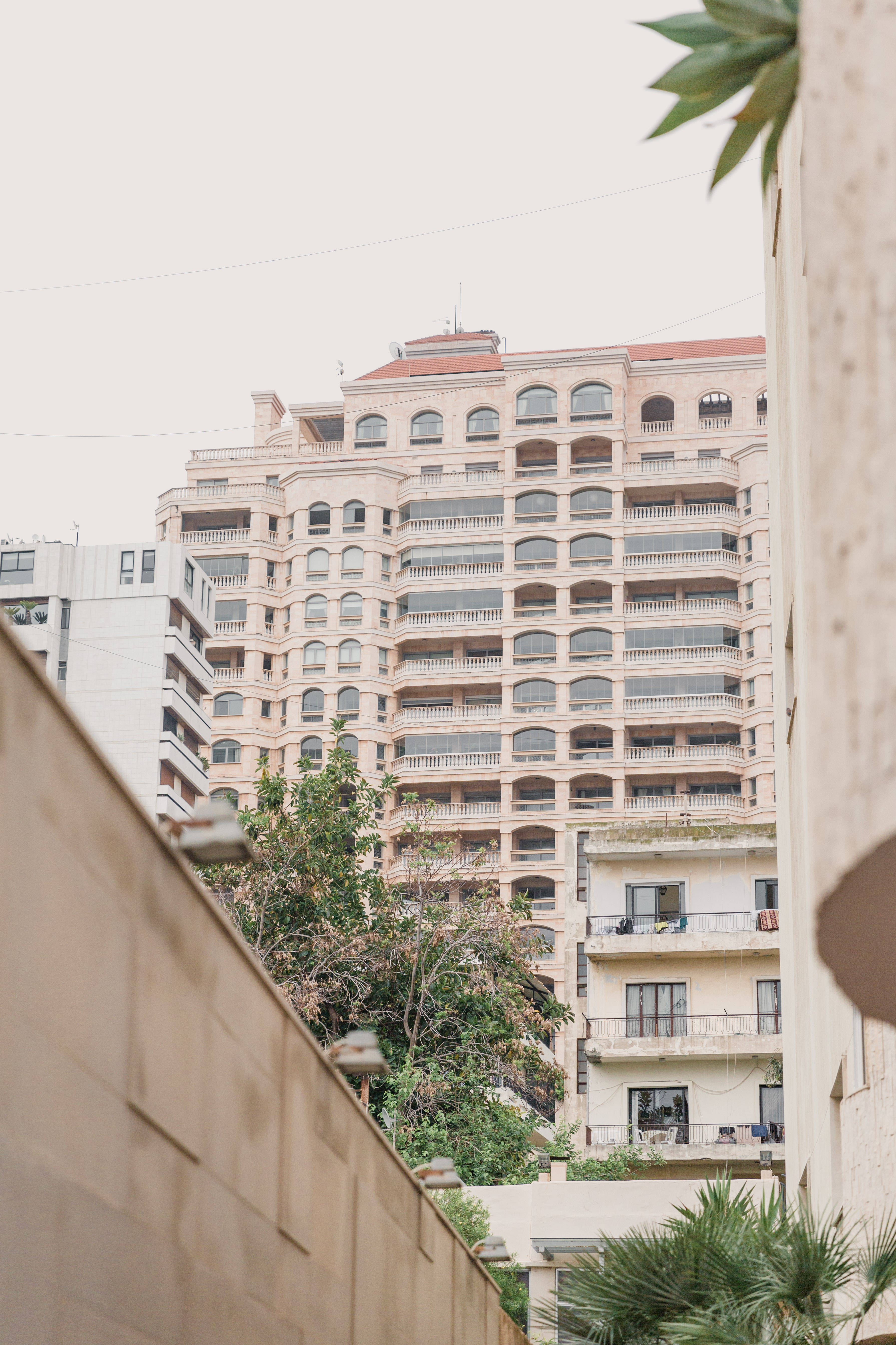 Lebanon 桌布 壁紙 — 一棟高大的米色公寓大樓，帶有拱形窗戶和陽台，聳立在狹窄的城市小巷之間，周圍環繞著綠植。