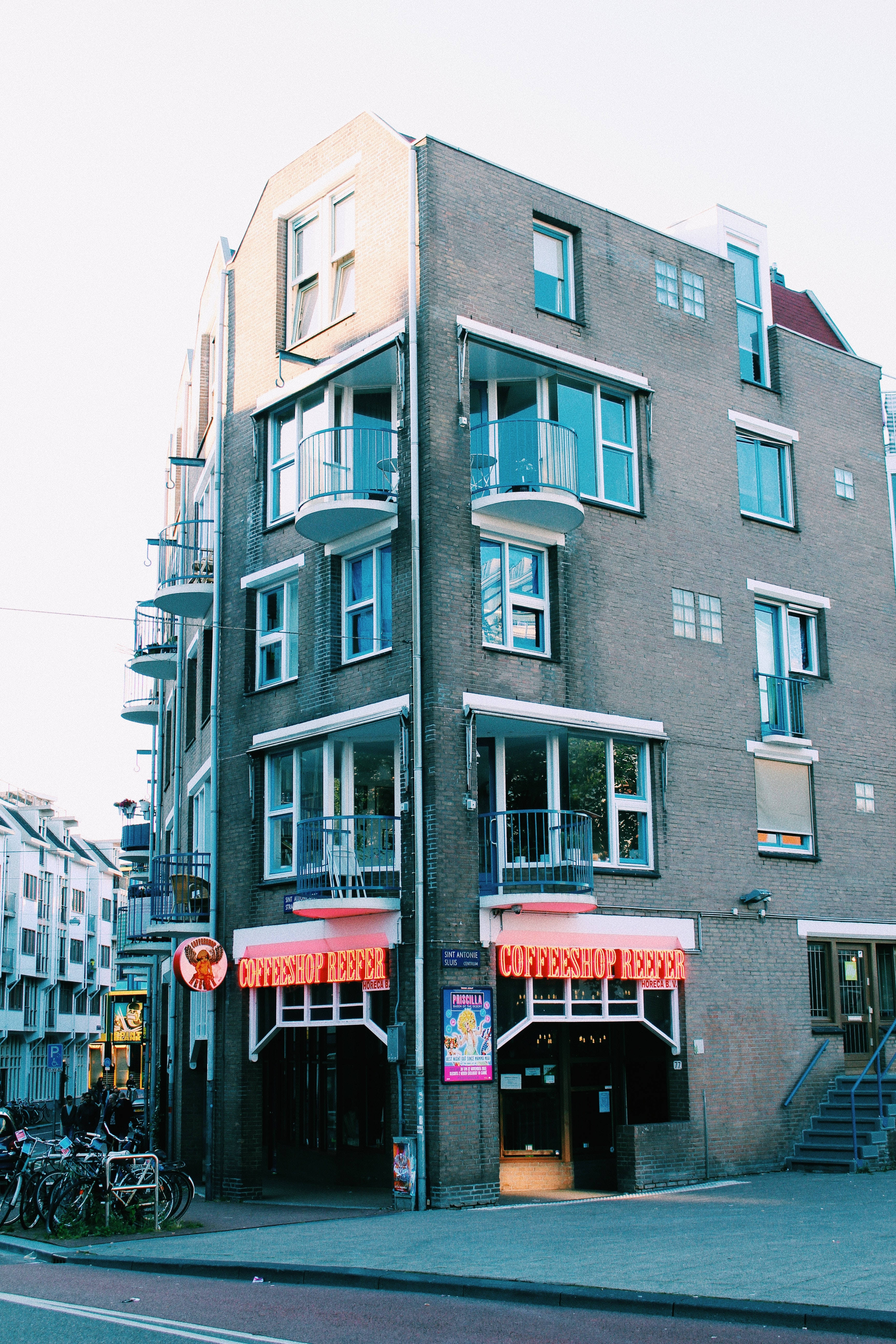 Coffee Shop 桌布 壁紙 — 一棟高層磚建築，帶有陽台，街邊有一間咖啡店，天空蒼白。